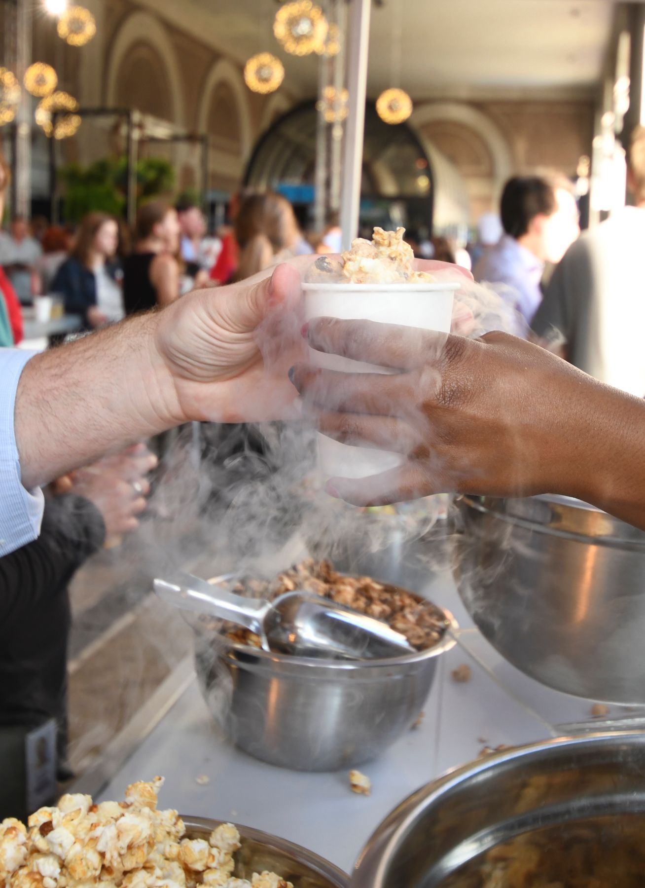 Hands passing a cup of smoky popcorn at a vendor's stall in an outdoor setting.
