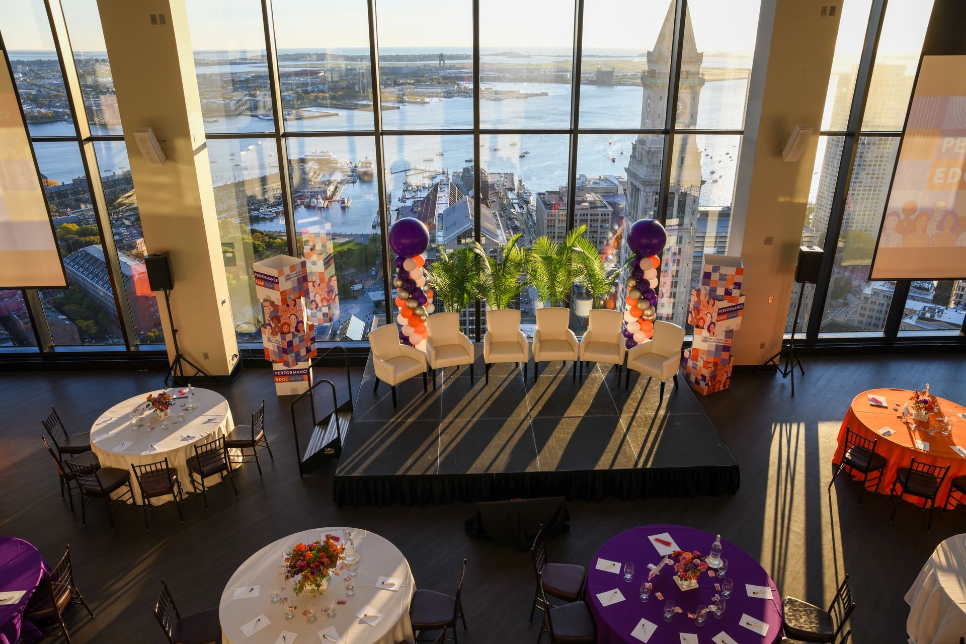 Event setup in a high-rise venue with city/water views. Round tables with centerpieces surround a stage with chairs.