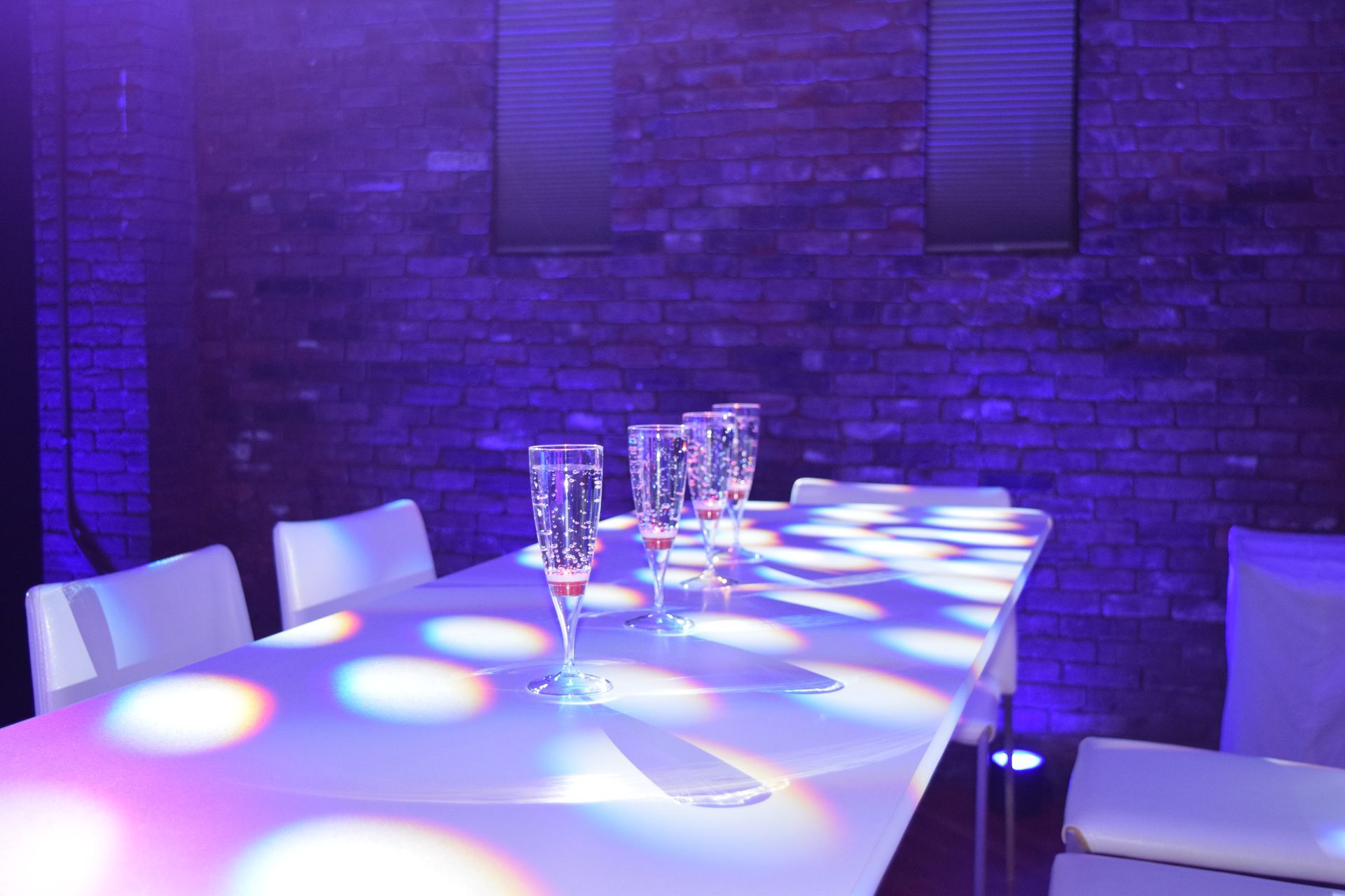 A table set with champagne glasses in a dim, purple-lit room. Brick wall backdrop and spotlight effect on the table.