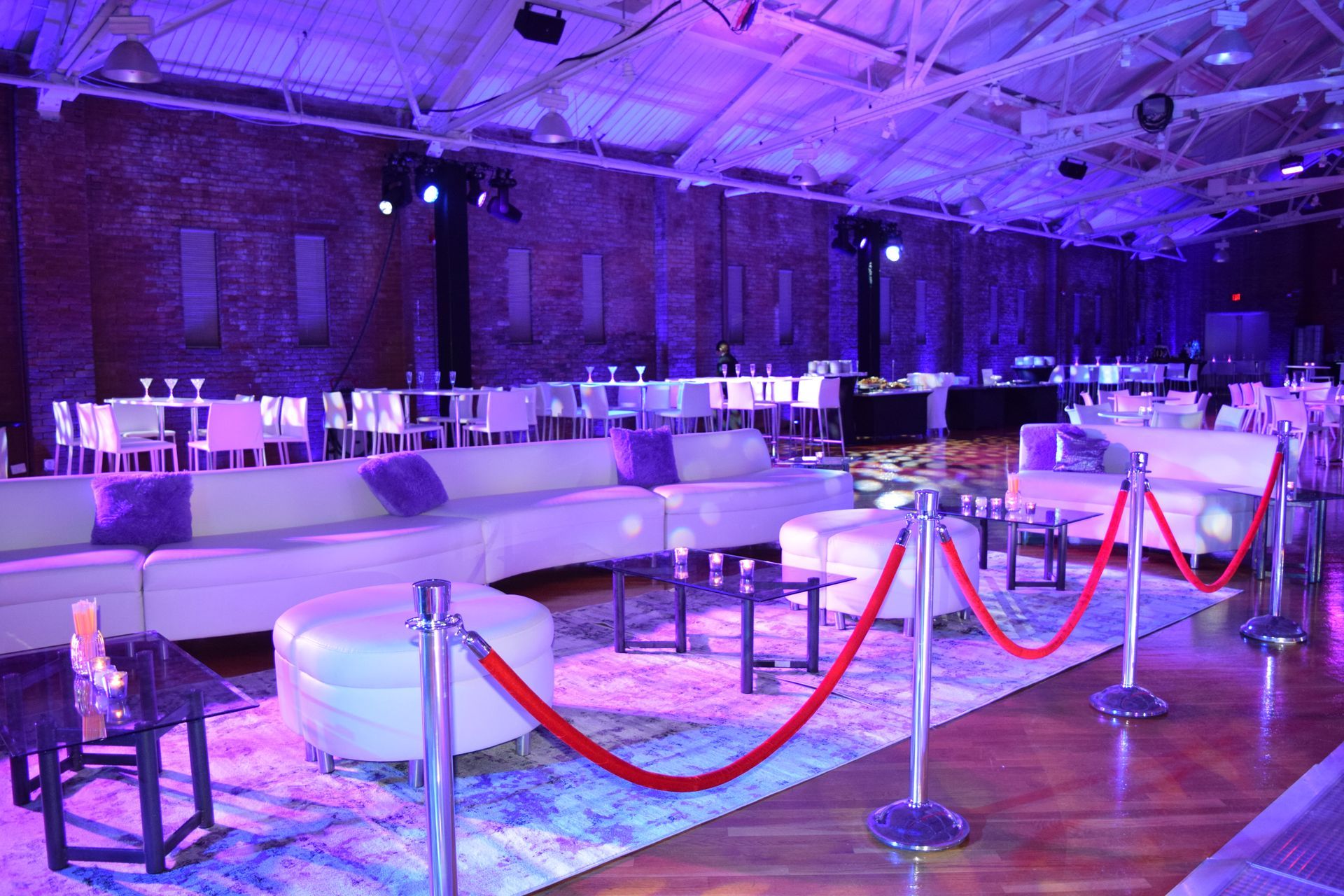 Event space with white couches, tables, red ropes, purple lighting, and exposed brick walls.