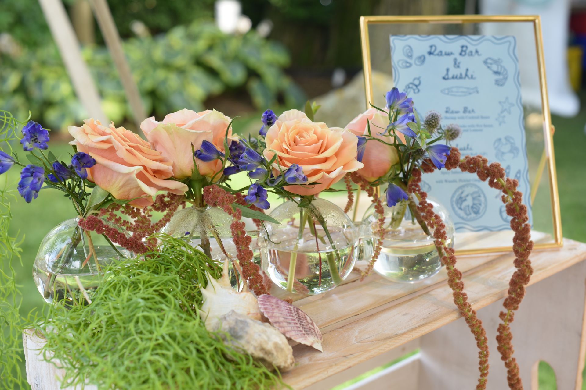 Floral arrangement: peach roses, blue flowers, and hanging brown amaranth in glass vases on a wooden crate.