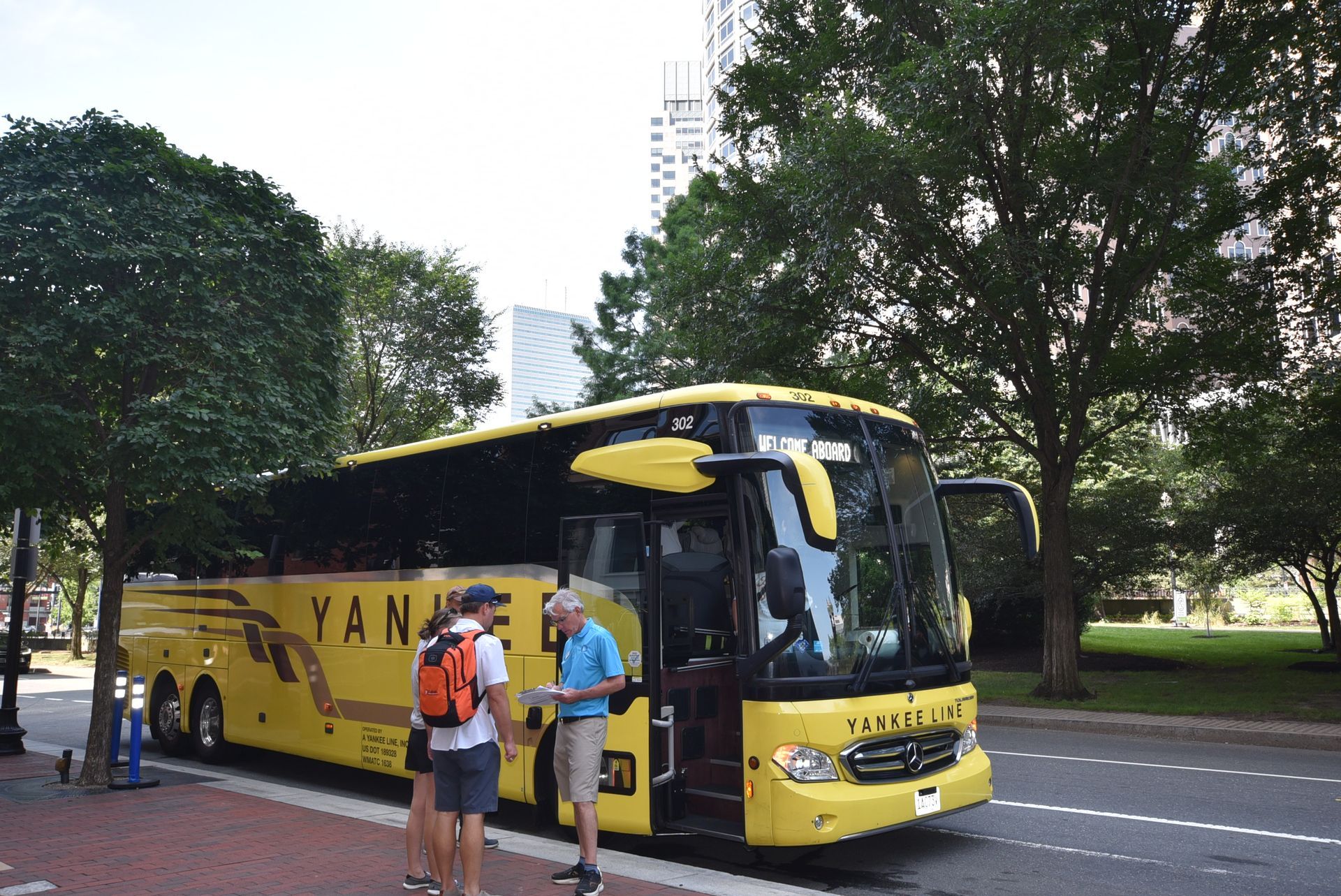 Yellow Yankee bus parked on a city street. Two people talking near the open door. Trees line the sidewalk.