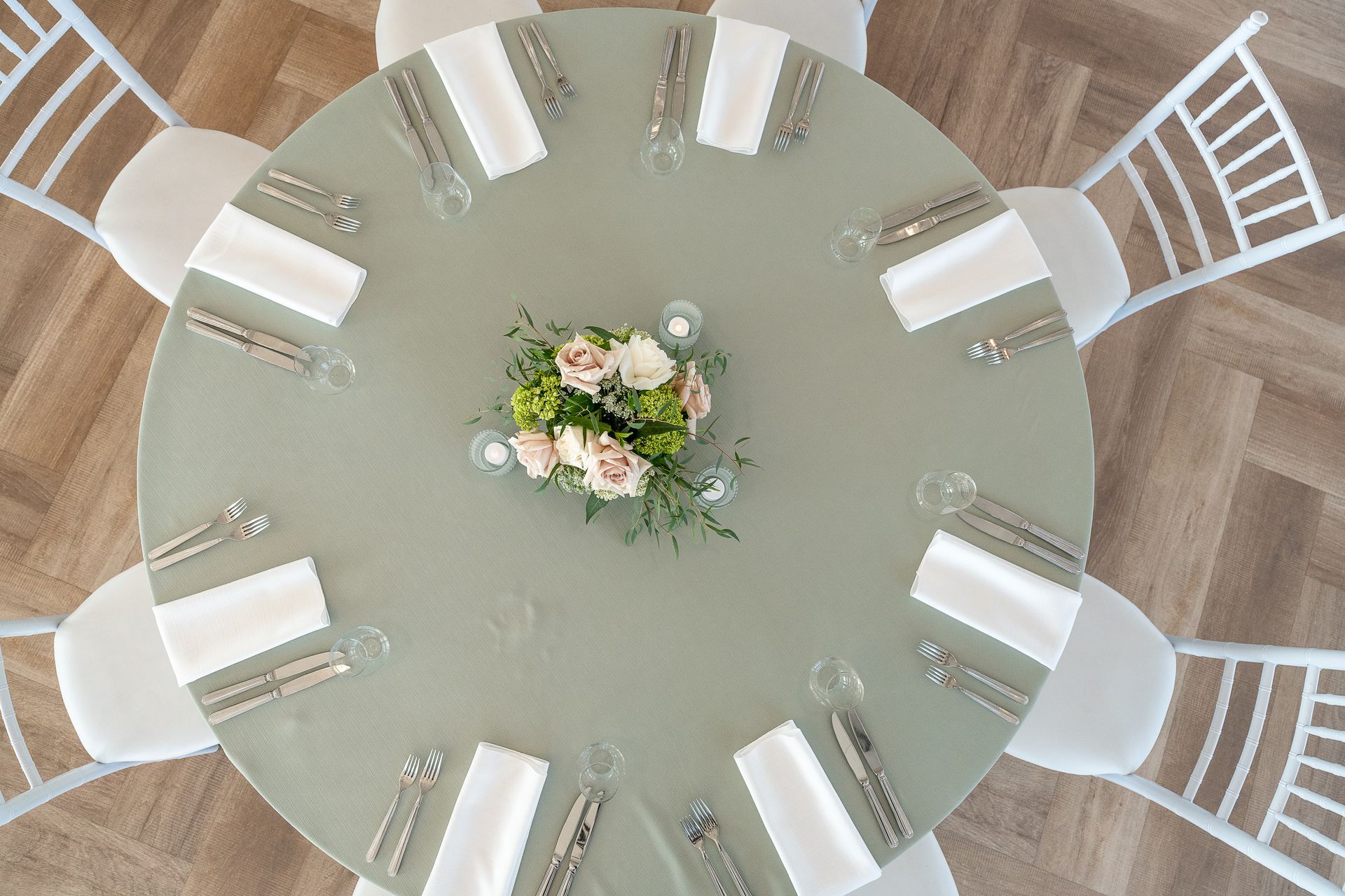 Round table set for a meal, with place settings and a floral centerpiece; surrounded by chairs.
