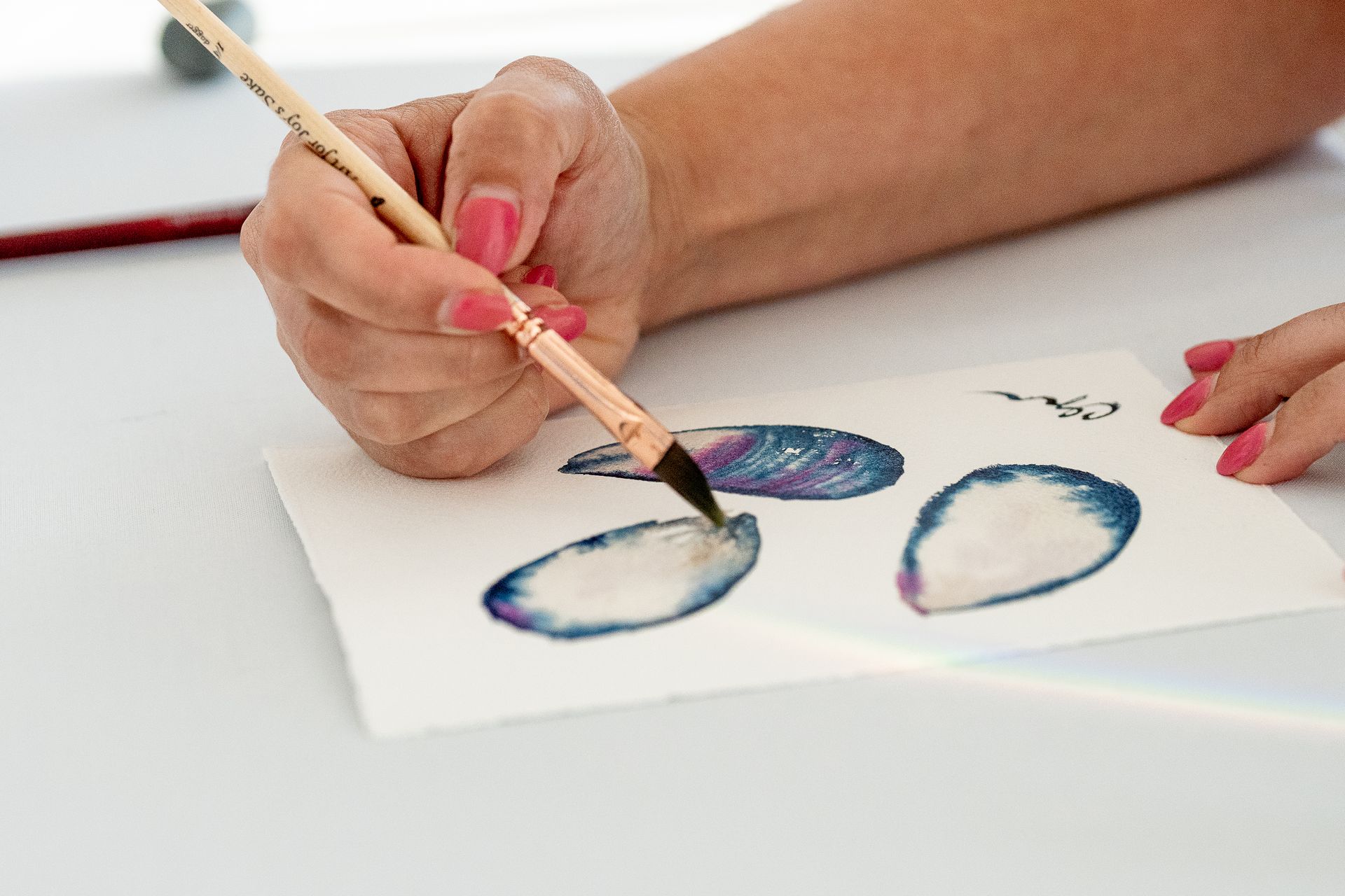 A person's hand painting blue watercolor shells with a brush on white paper, set on a white surface.