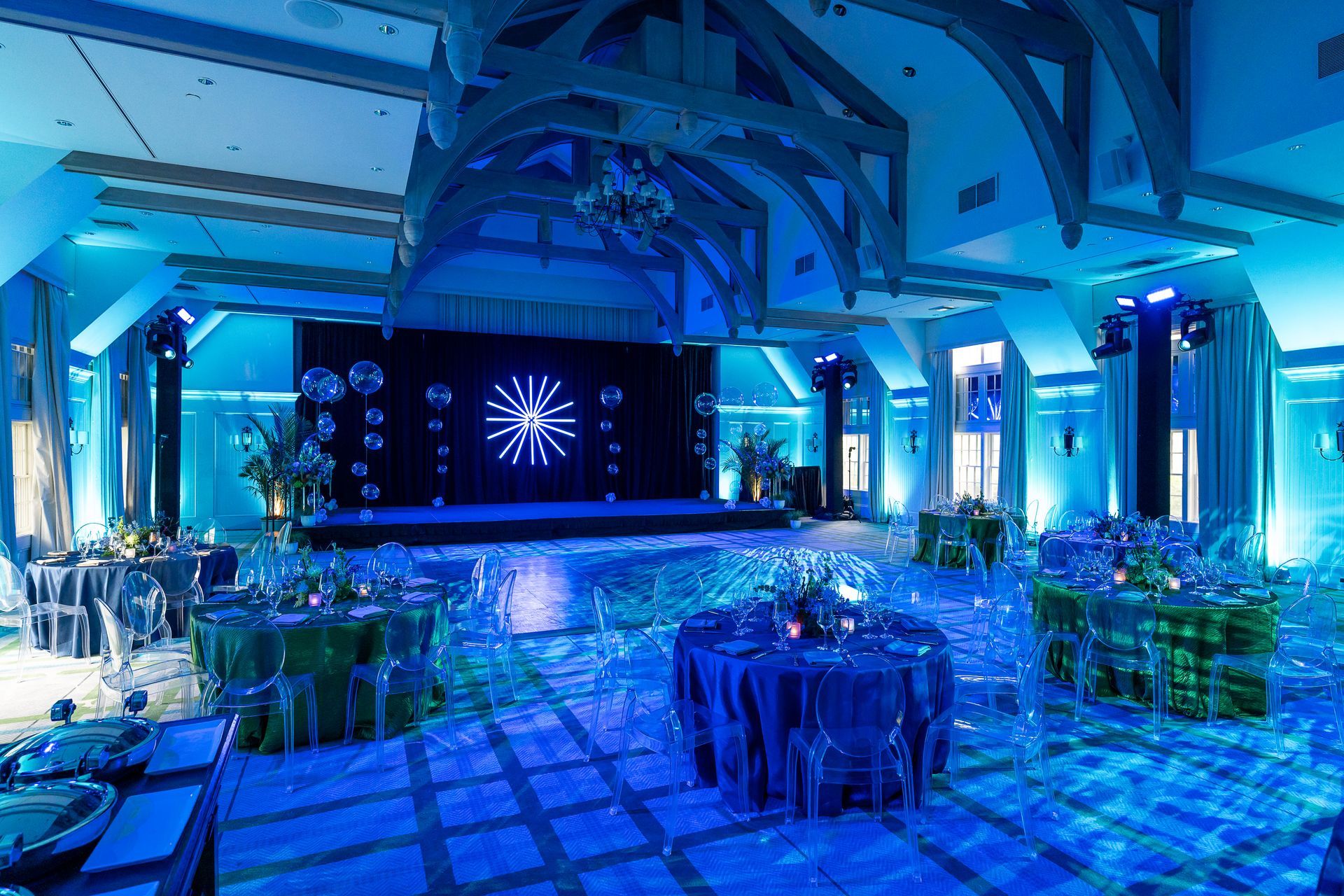 A blue-lit ballroom with tables, stage, disco balls, and ornate ceiling for an event.
