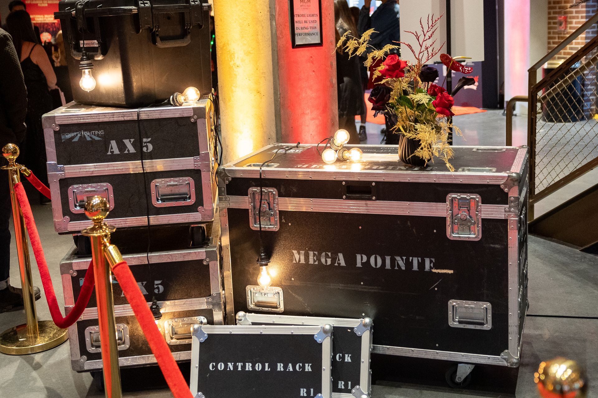 Stage equipment stacked with flowers, lit by Edison bulbs; gold rope barrier.