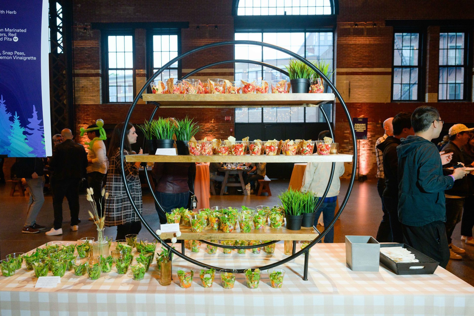 Food display at an event, with tiered circular shelves of packaged snacks and plants on a checked tablecloth.