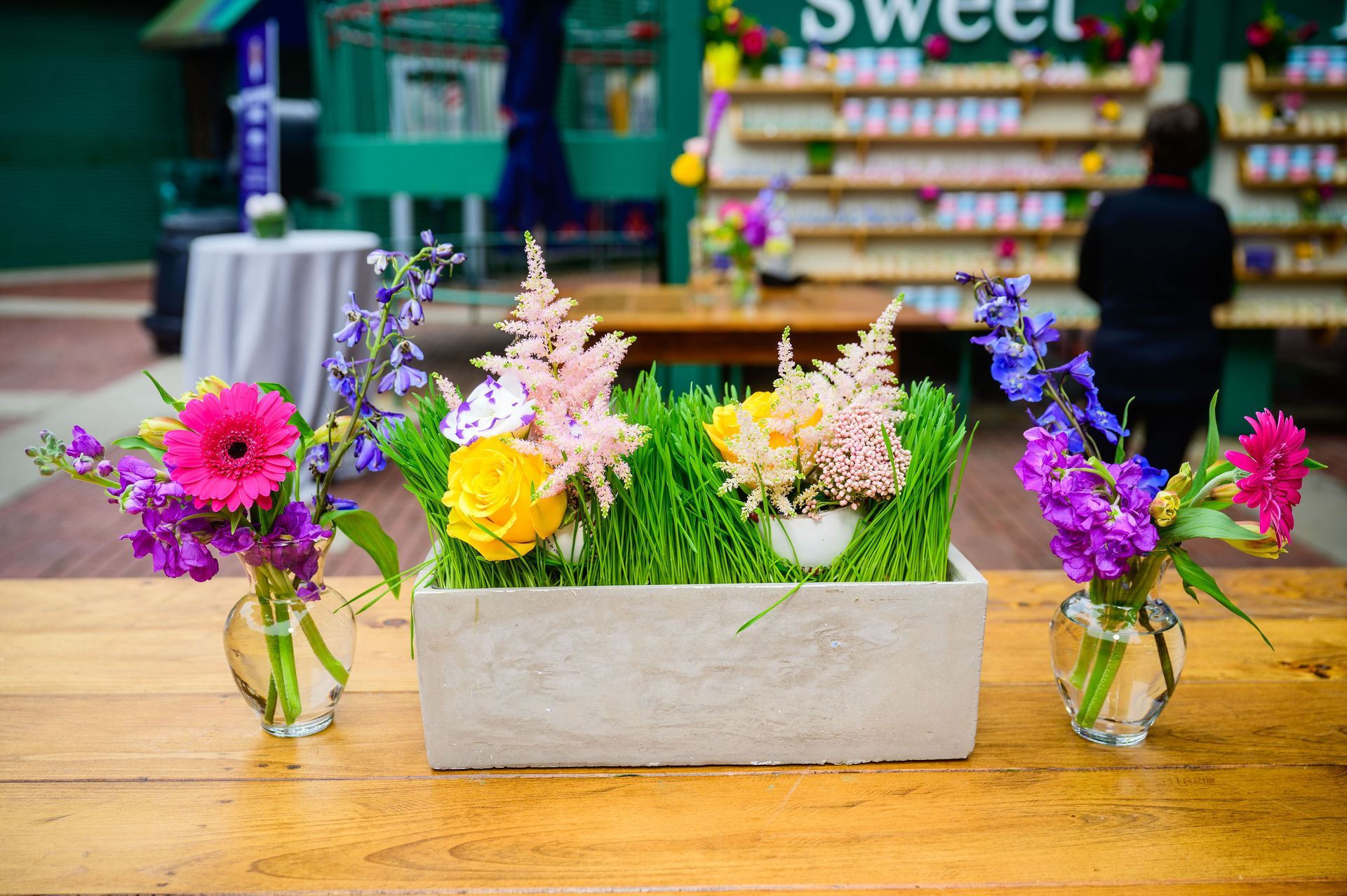 Floral arrangement on a wooden table: pink, yellow, and purple flowers in glass vases and a rectangular planter with green grass.