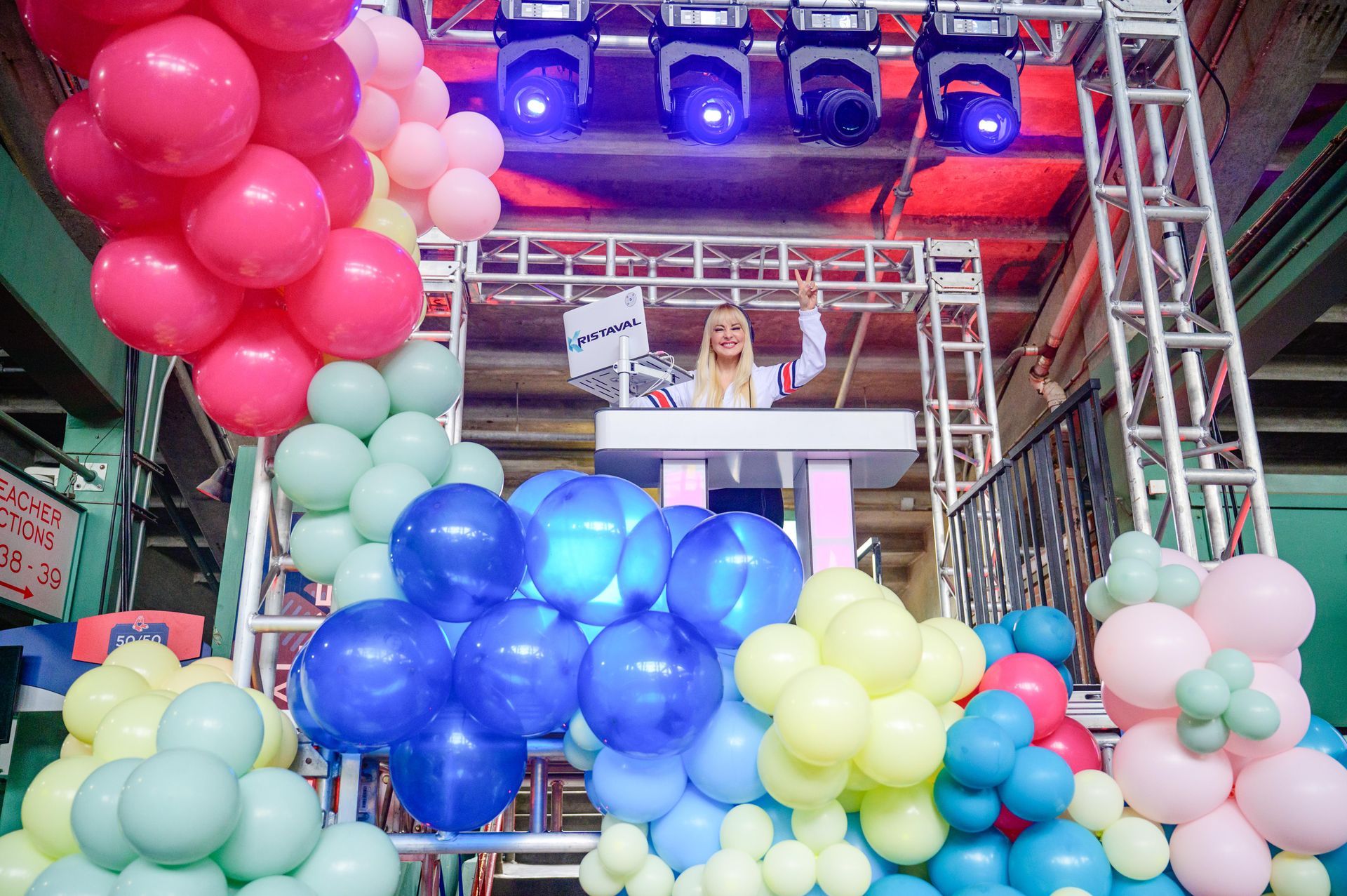 Blonde DJ at a colorful event with balloons and stage lights, waving from a podium.
