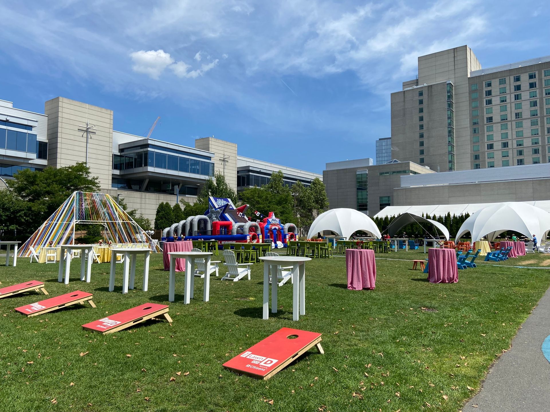 Outdoor party with games, tables, tents, and a bouncy house on a grassy lawn in front of buildings under a blue sky.