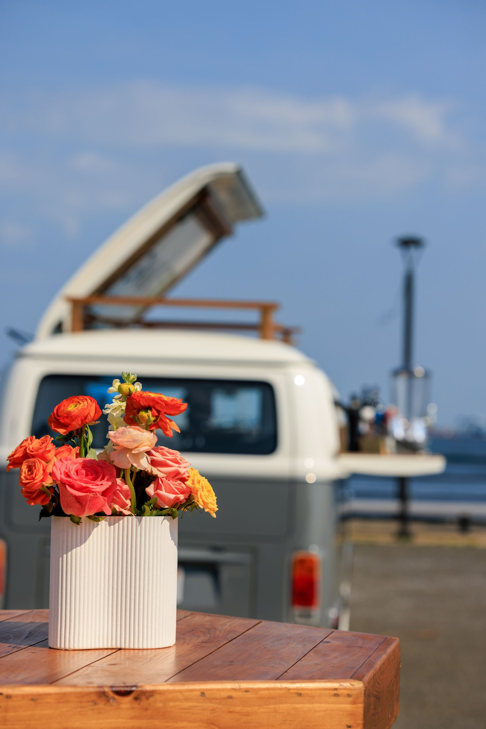 Flowers in white vase on a wooden table, with a vintage campervan in the background.