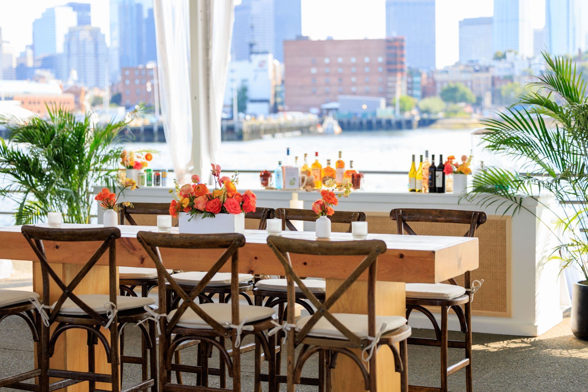 Outdoor dining area with wooden table, chairs, and bar, flowers, and city skyline view.