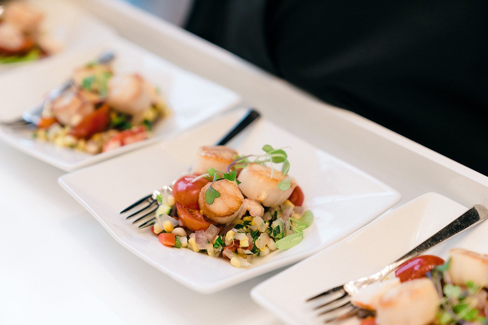 Plated seared scallops with corn, tomatoes, and greens. White plates on a tray.