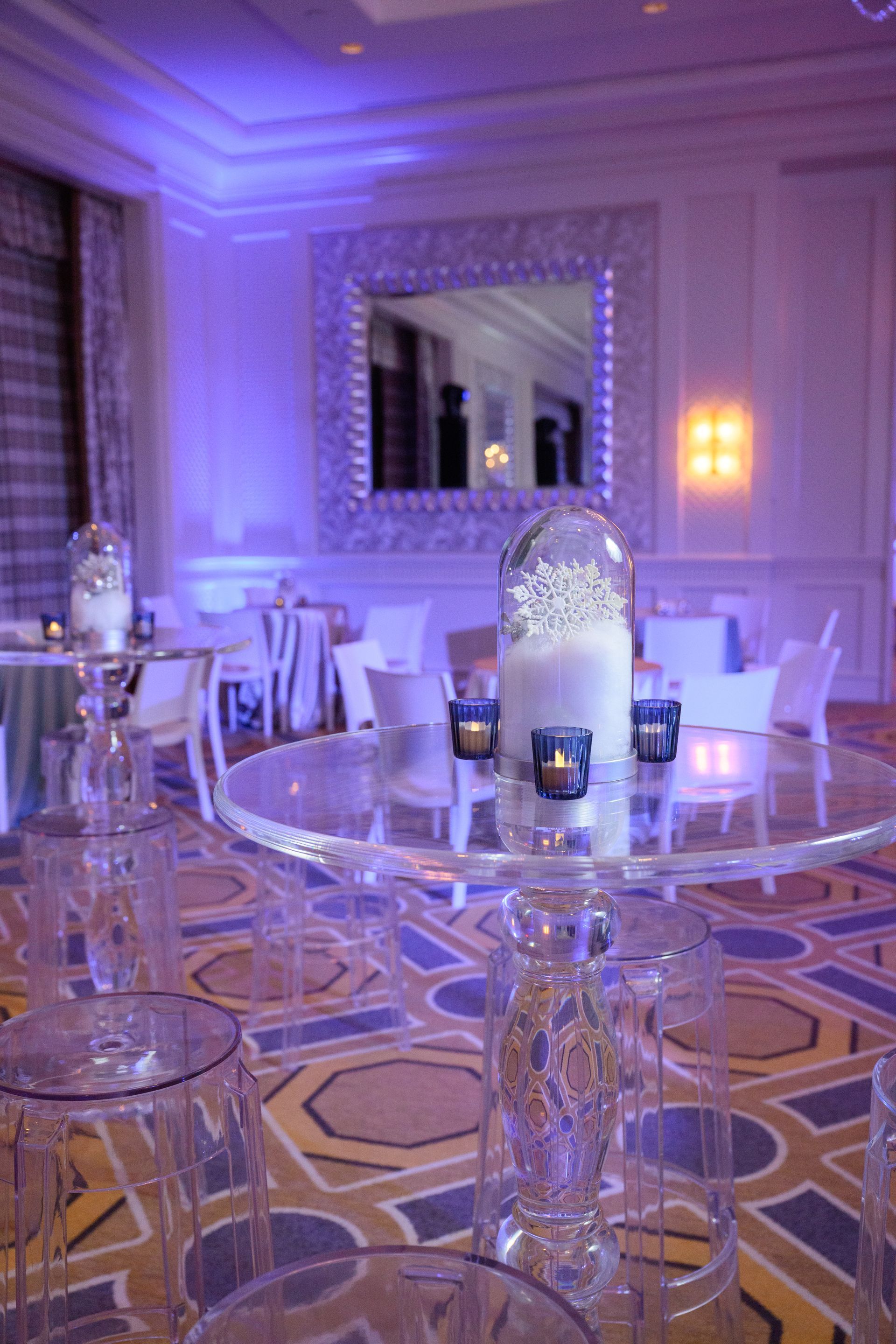 Elegant venue with translucent tables, white chairs, and purple lighting, set for an event.