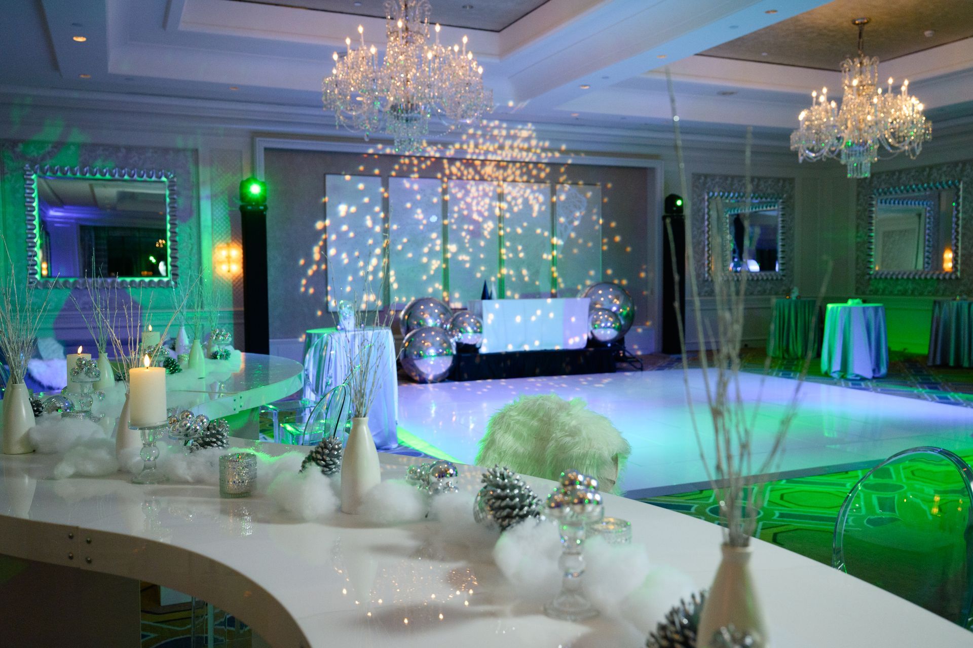 A decorated ballroom with white and silver decor; mirrored walls, chandeliers, and a white dance floor.