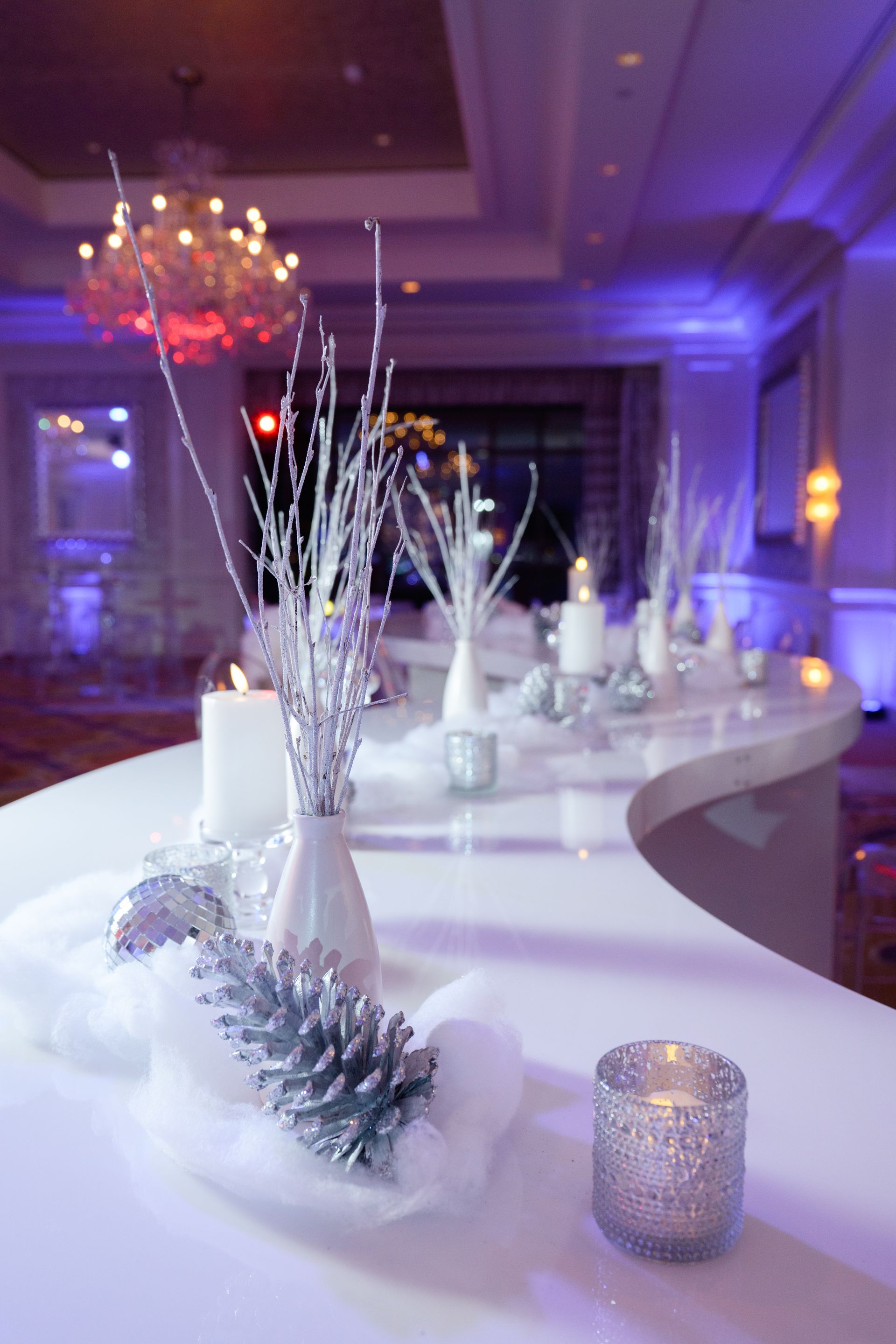 Winter-themed table centerpiece with candles, silver ornaments, and frosted branches, in a room with purple lighting.