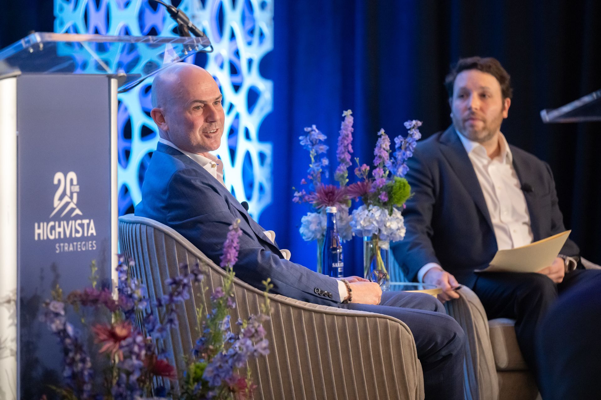 Two men in suits on stage at conference. One speaks, the other listens with paper in hand.
