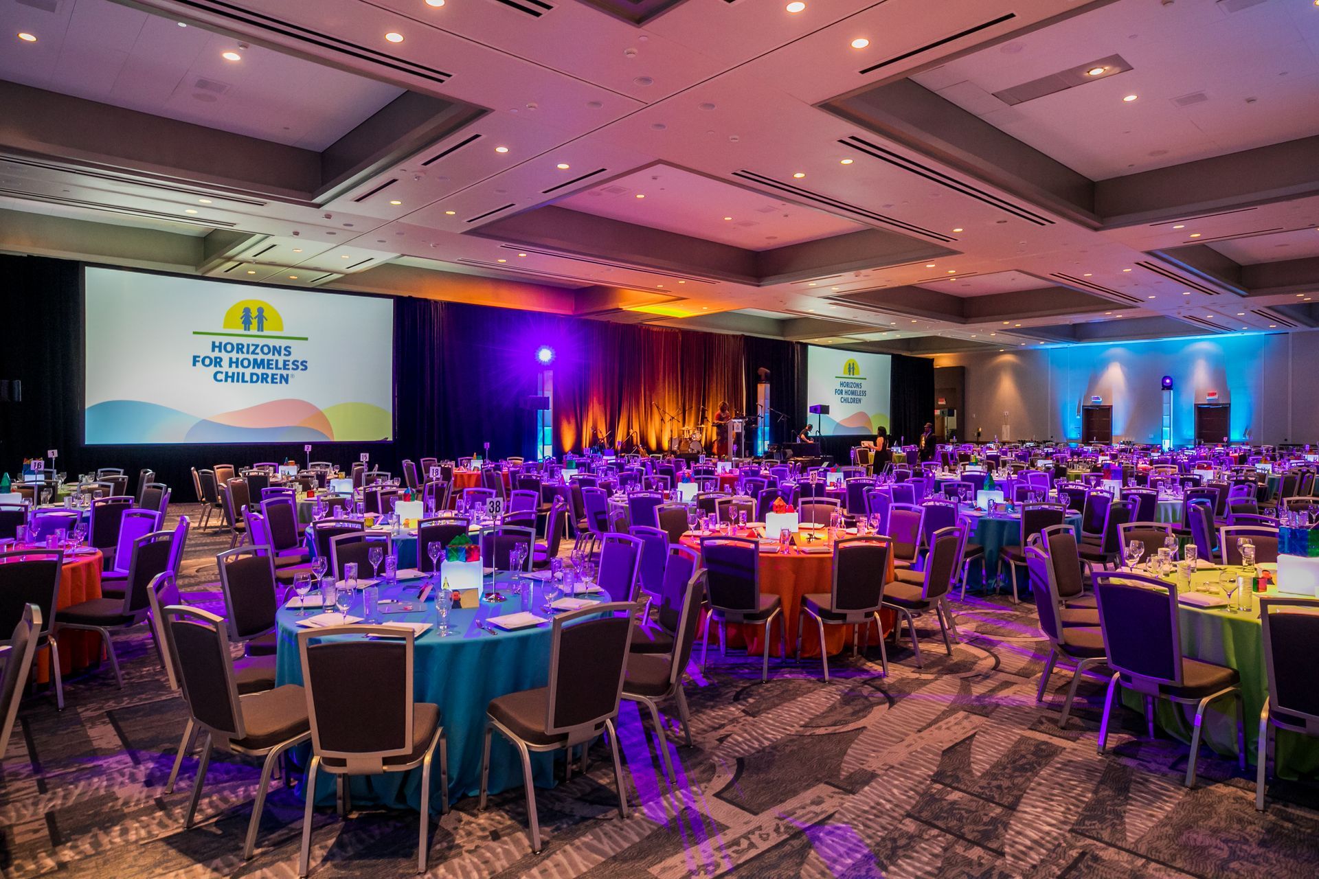 Large banquet hall with tables set for a colorful event, screens, and stage.