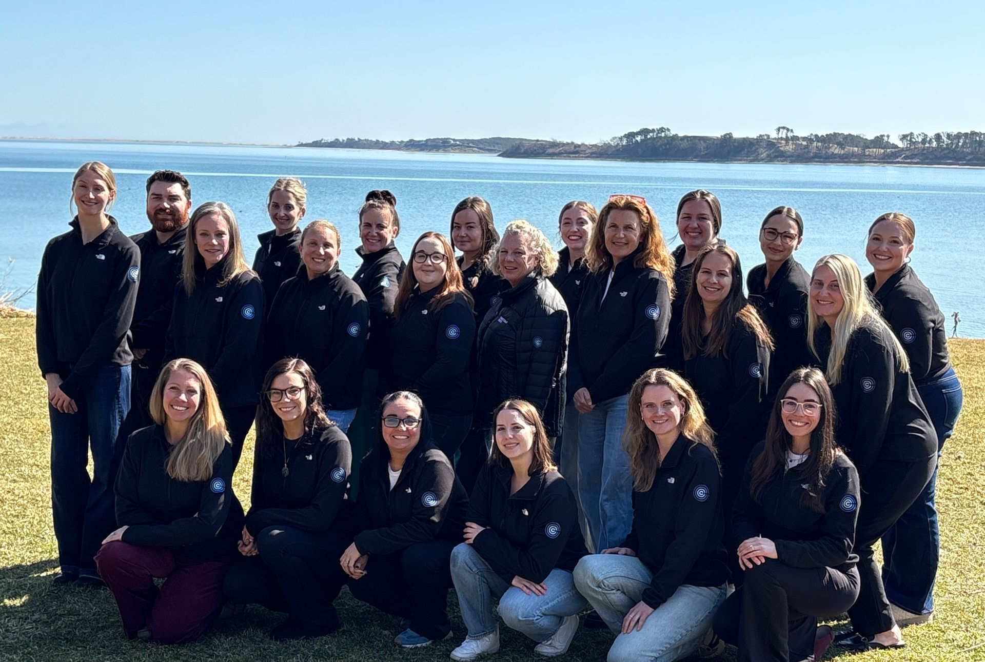 A large group of people in matching dark jackets stand on a grassy lawn by the water under a clear blue sky.