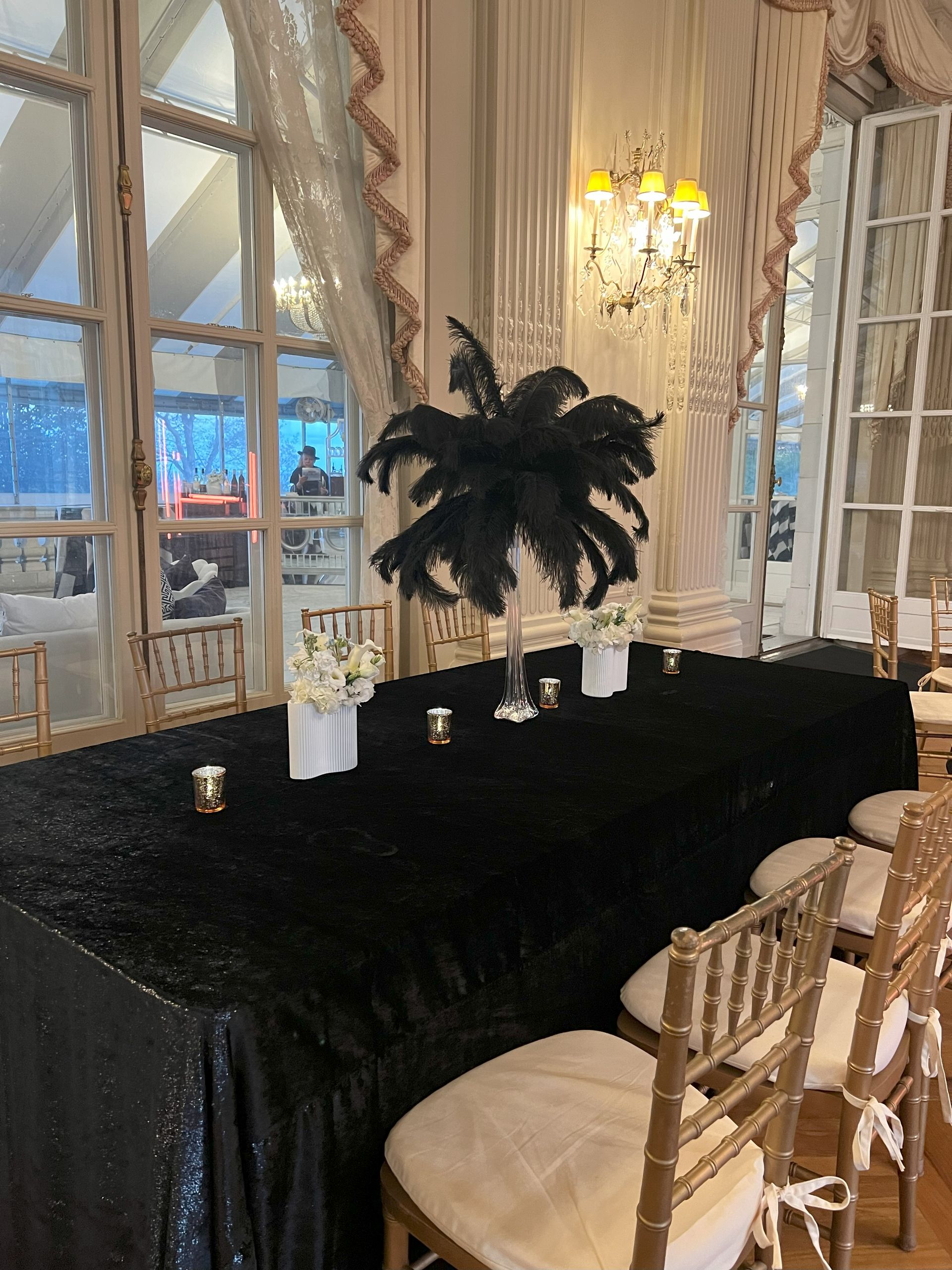 Elegant table with black sequin tablecloth, feathered centerpiece, gold chairs, and large windows.