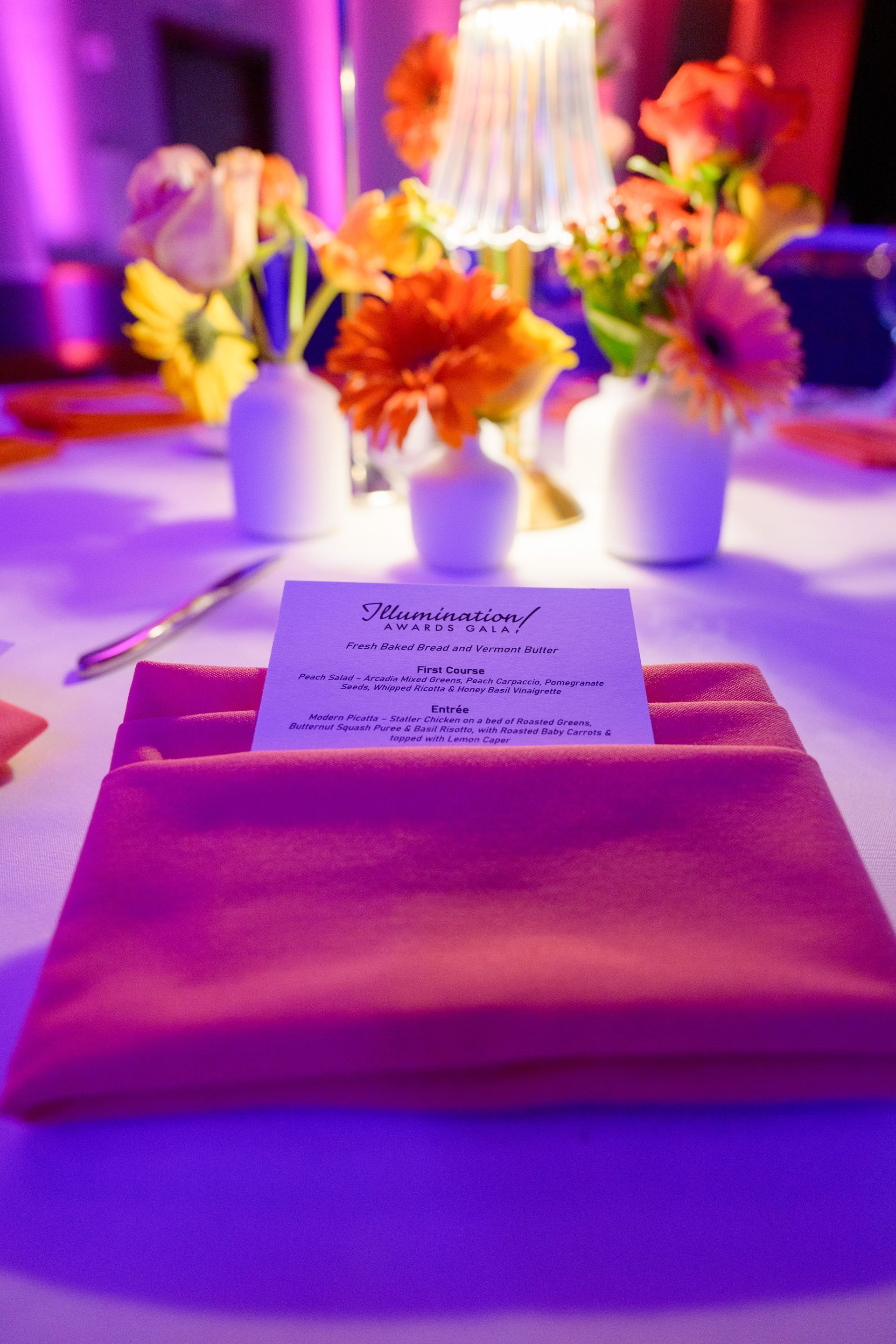 Table setting with orange napkin and floral centerpieces under a lamp.