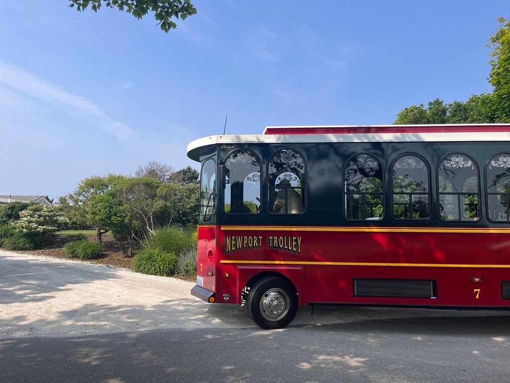 Red trolley with black trim parked on a gravel road next to greenery, under a blue sky.