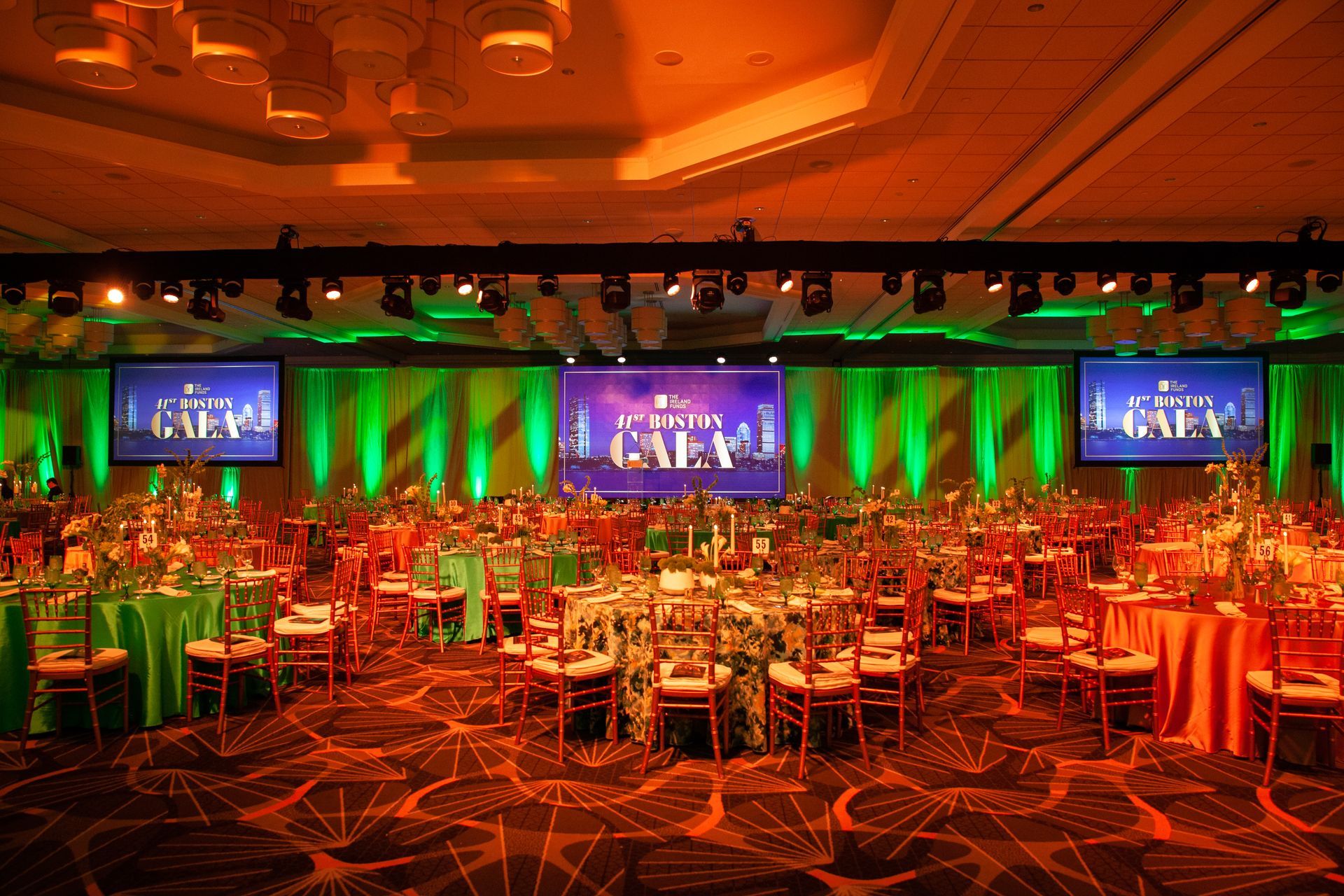 Ballroom set for gala, tables with colorful linens. Stage with screens, green and orange lighting.