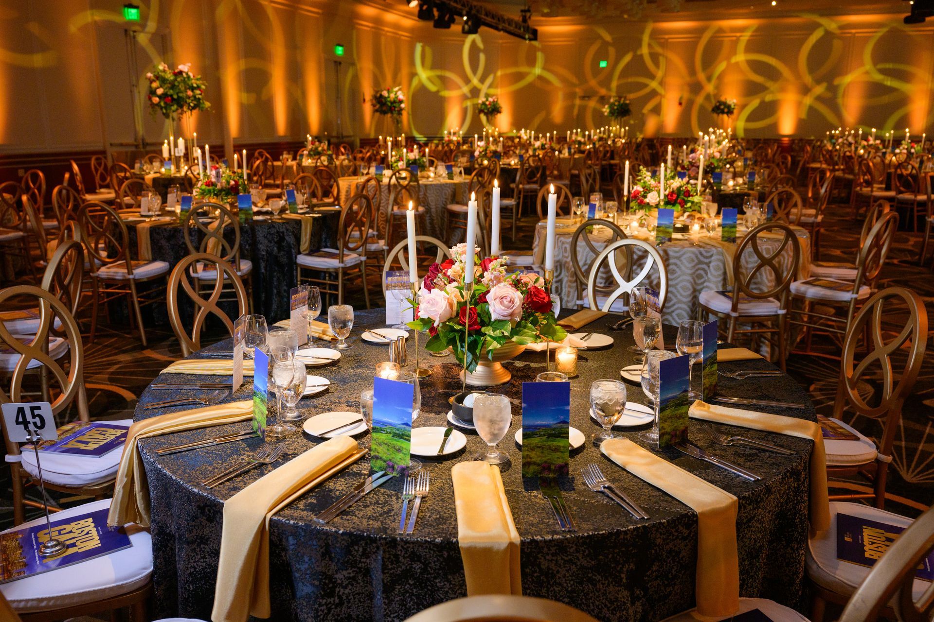 Formal banquet hall with round tables, candles, and floral centerpieces. Gold and black decor.