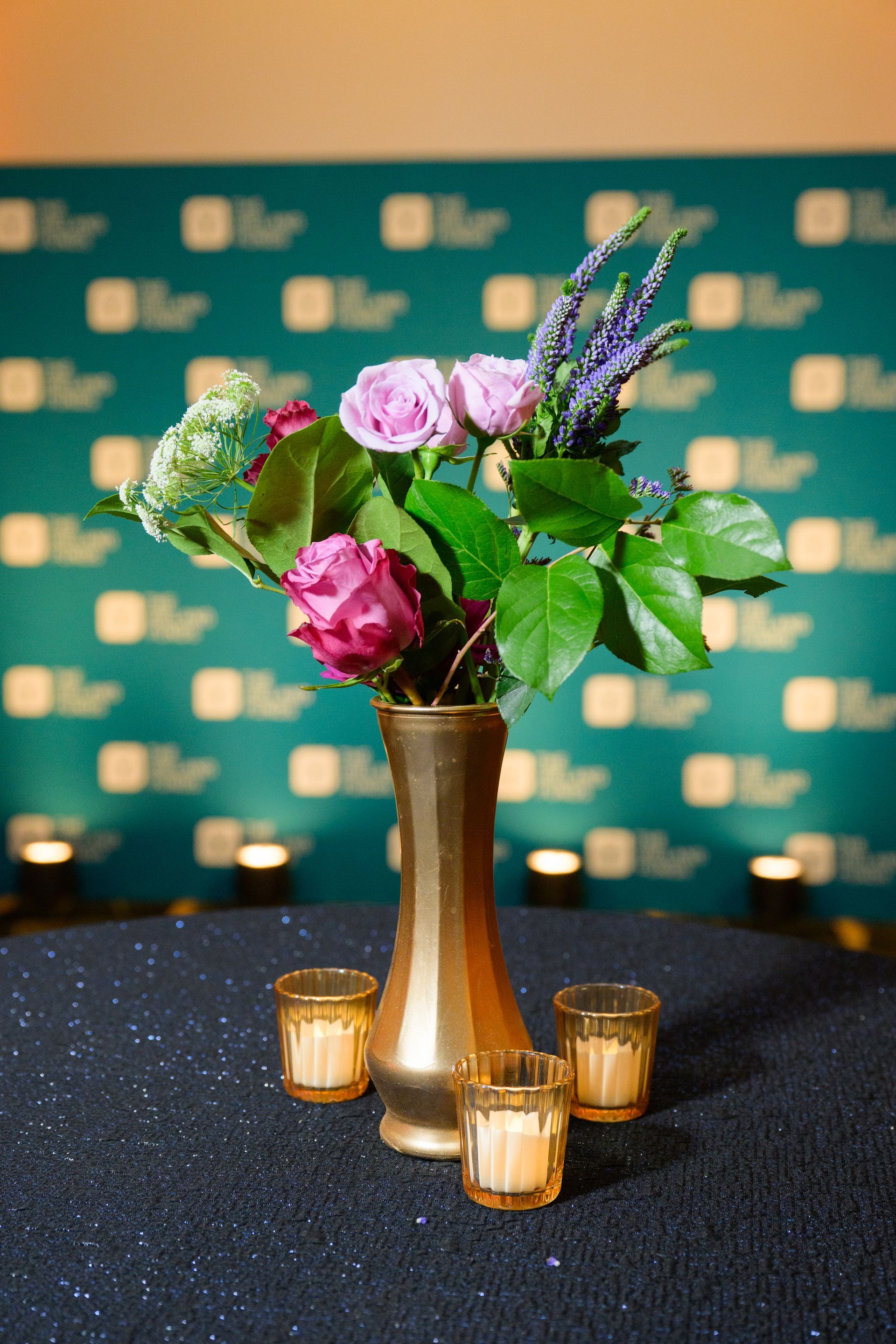 Floral arrangement in gold vase, with three candle holders on black tablecloth. Teal wall in background.