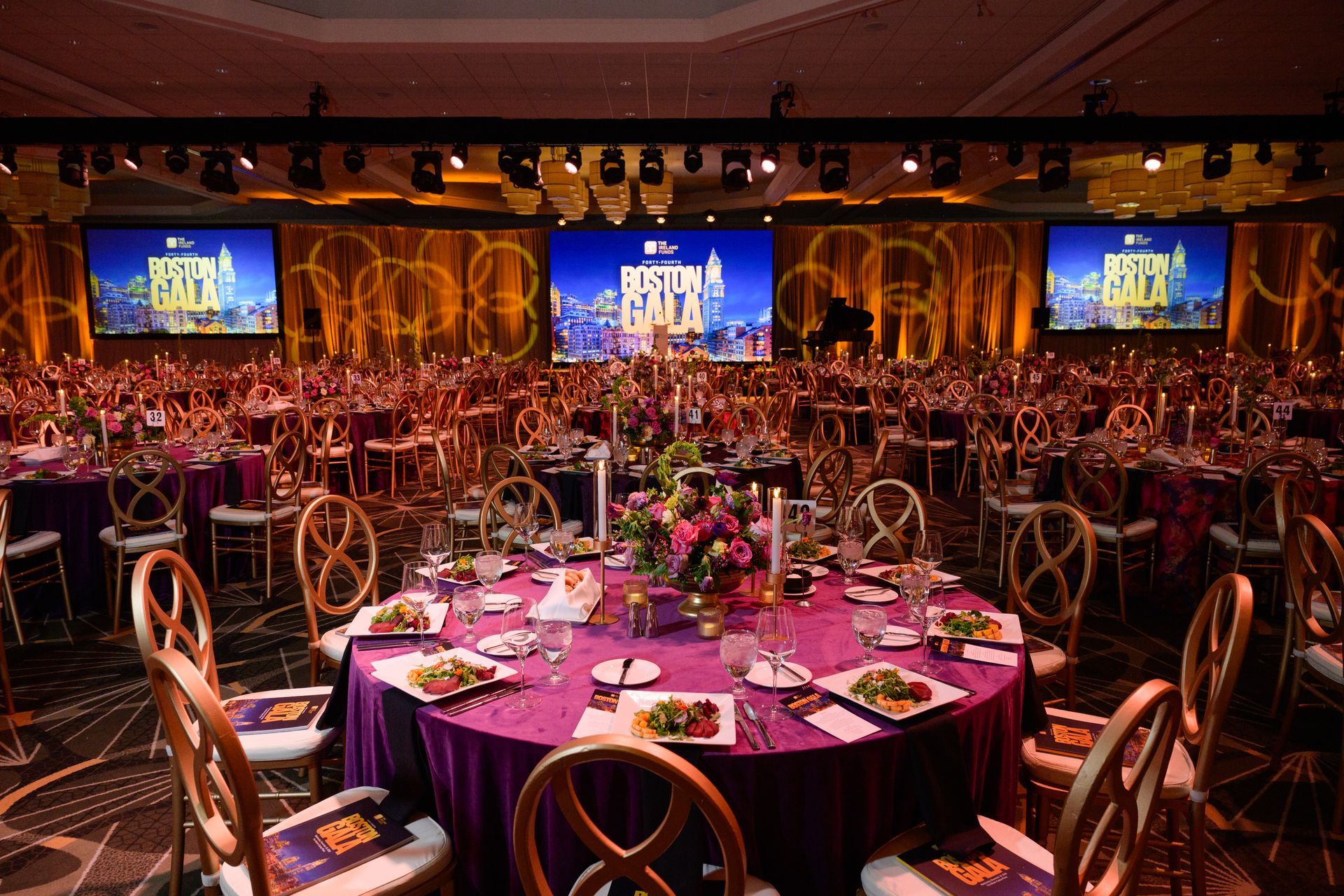Elegant ballroom set for a gala, with round tables draped in purple, gold chairs, and large screens.