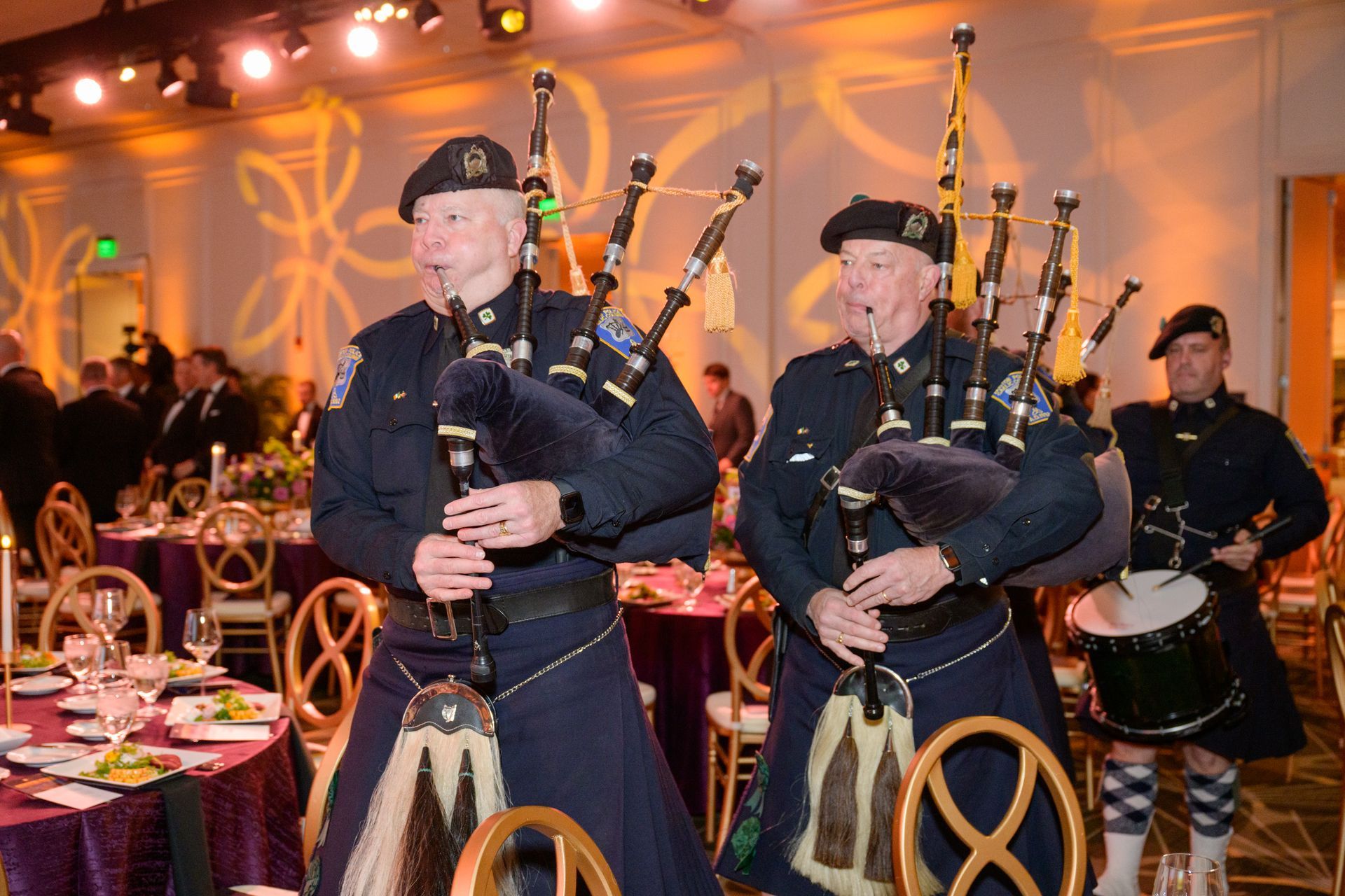 Two bagpipers and a drummer in uniform performing at a formal event, with tables and guests in the background.