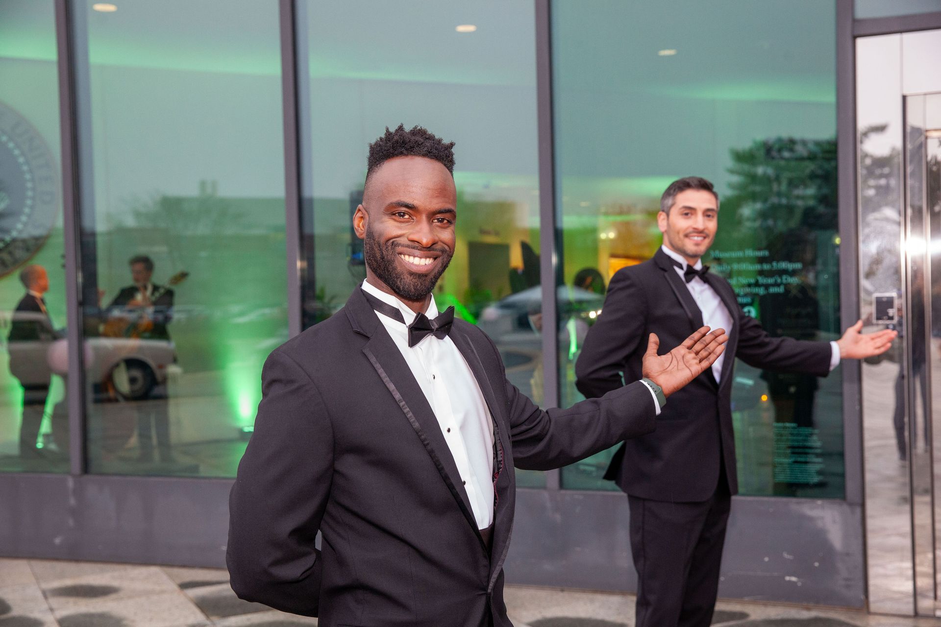 Two men in tuxedos welcoming guests at a building entrance. Green lighting and a band are visible.
