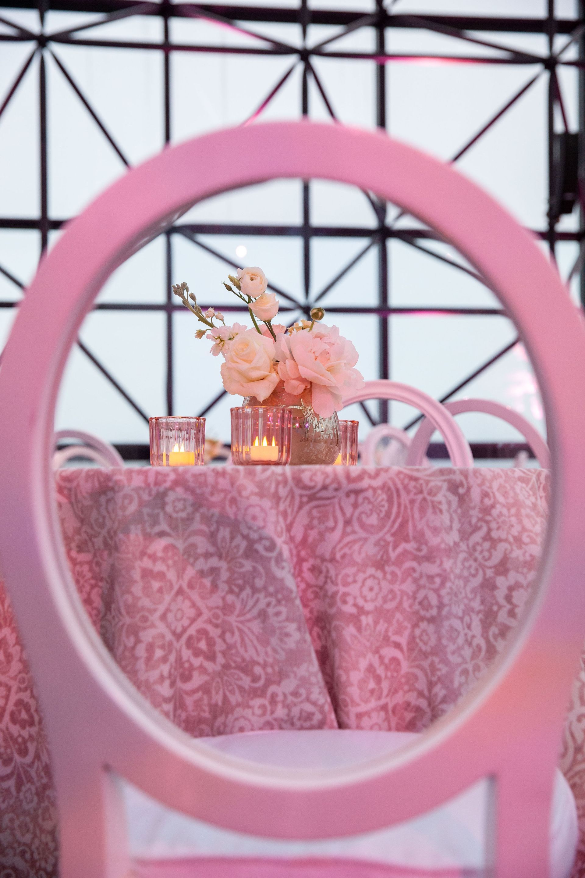 Pink table setting with flowers, candles, and patterned tablecloth seen through a pink chair.