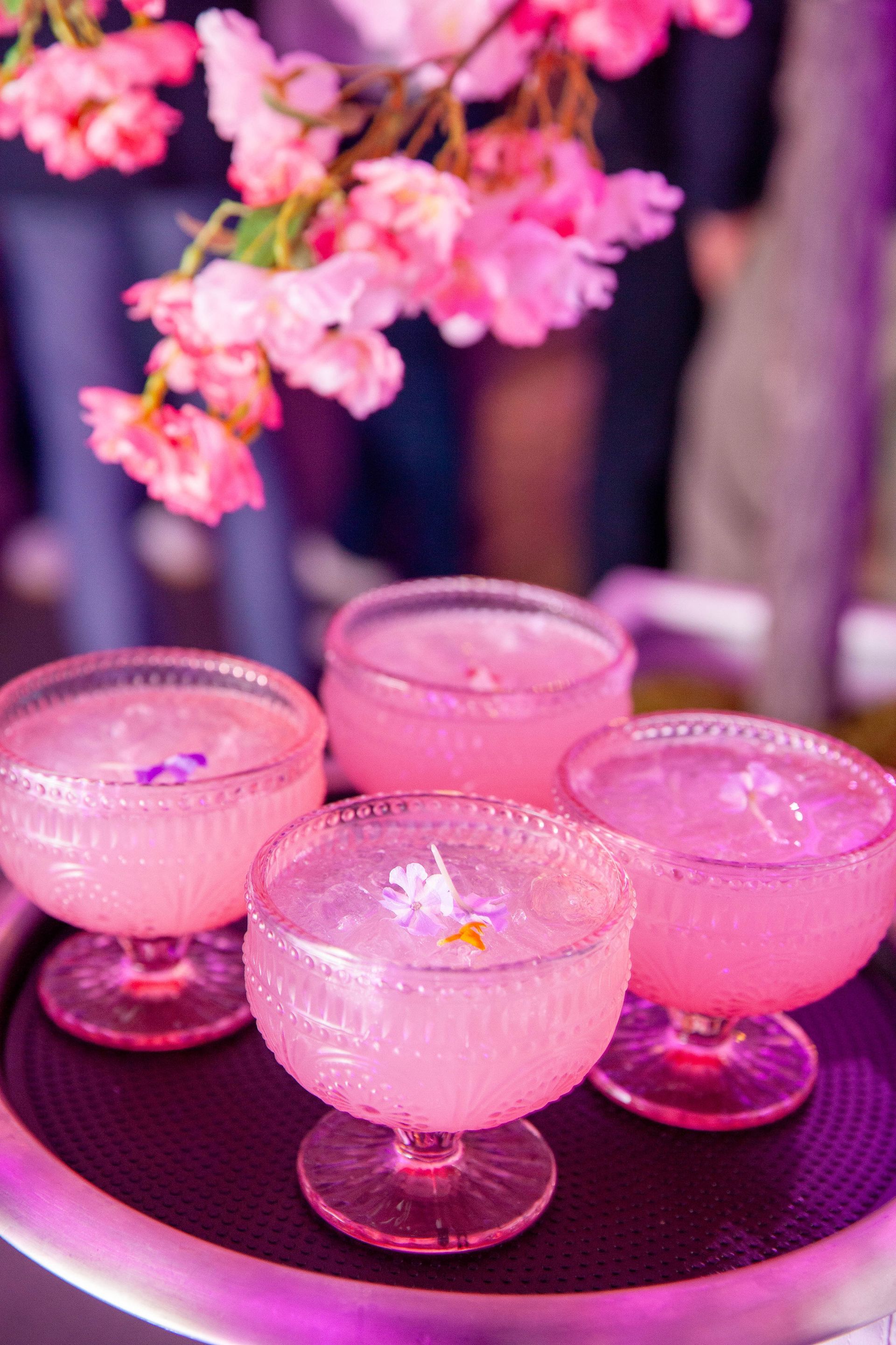 Four pink cocktails in glass bowls on a tray, decorated with flowers, under pink cherry blossoms.