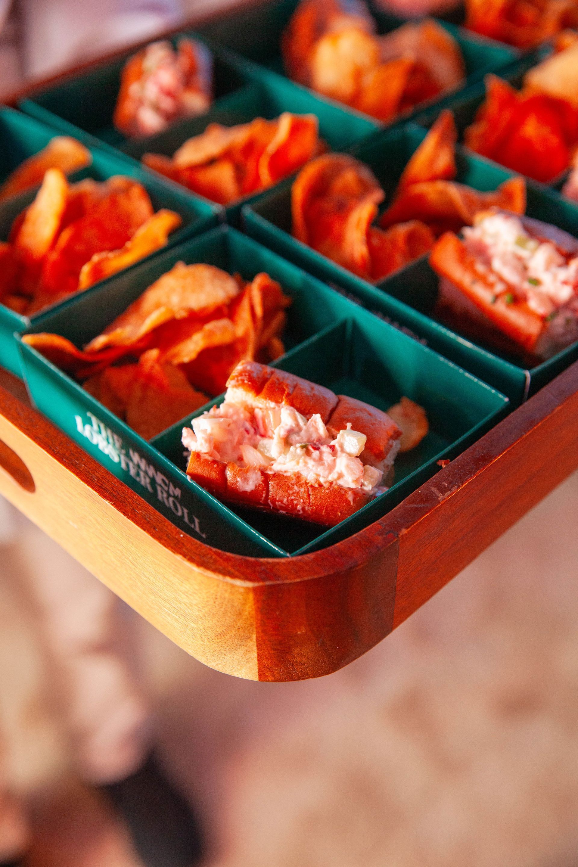 Wooden tray with various appetizers: lobster rolls, chips, in turquoise compartments.