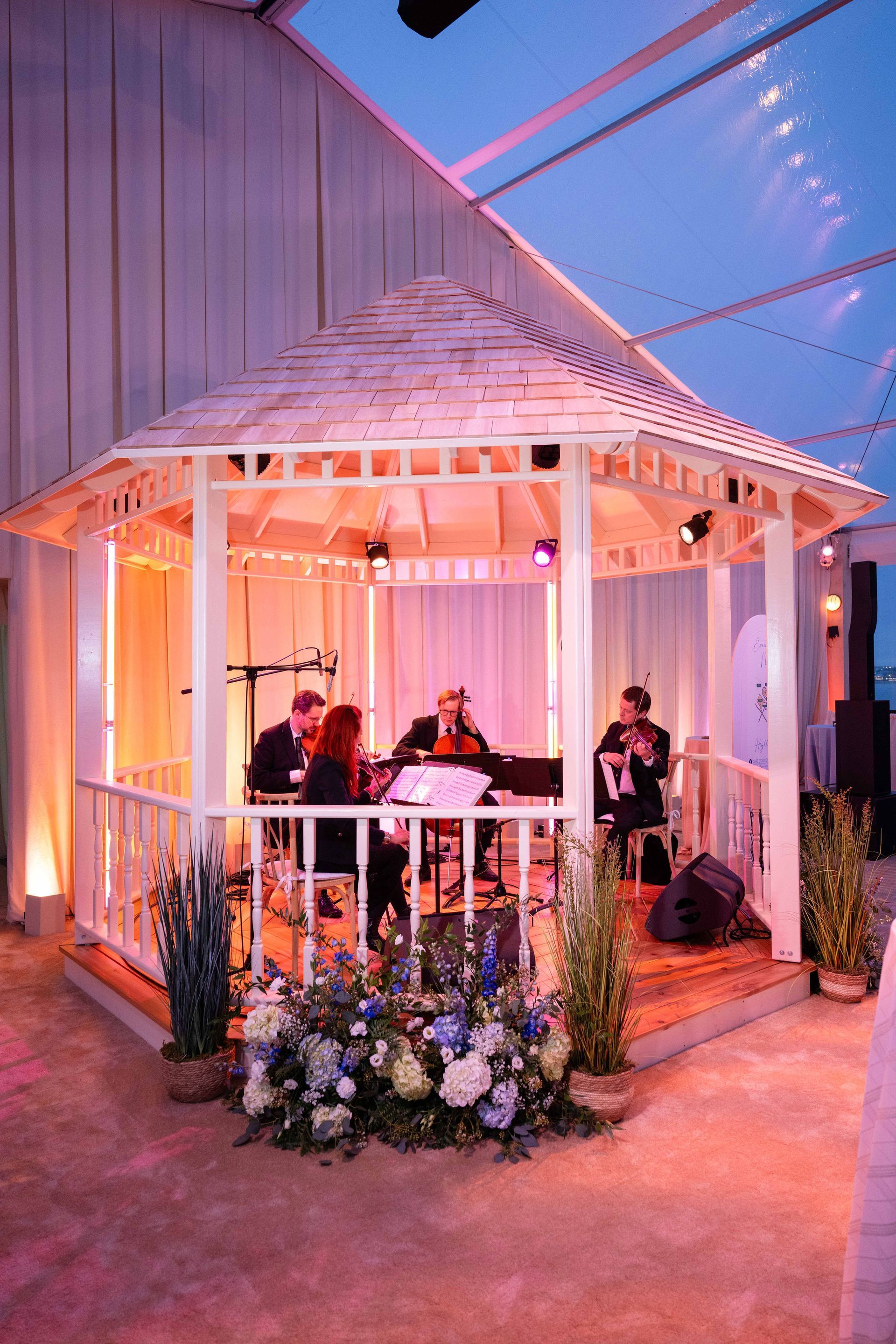 String quartet performs in a white gazebo decorated with flowers, under soft pink lighting.
