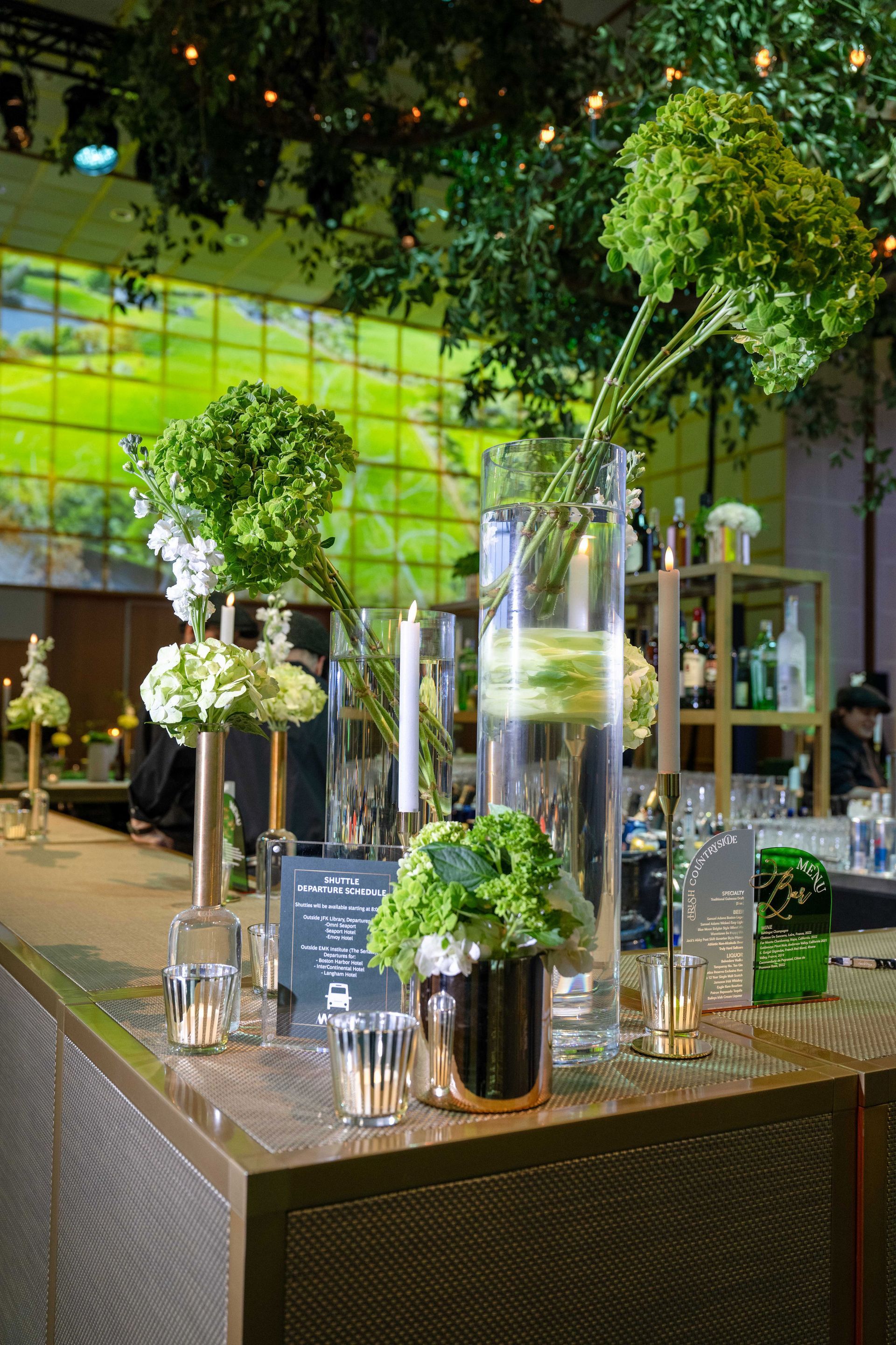 Bar with floral arrangements in vases, lit by candles and overhead lights. Green and white flowers.