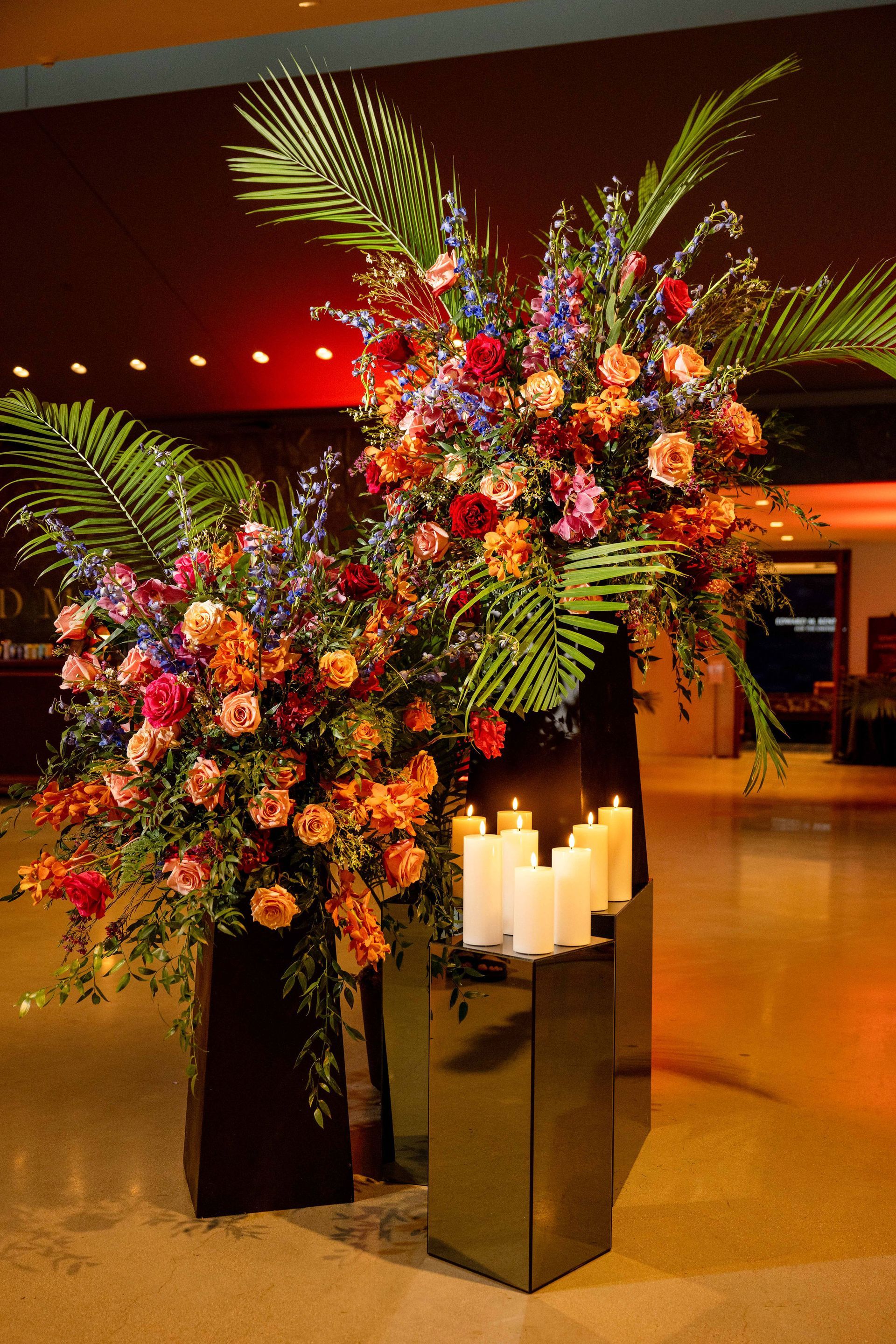 Floral arrangement with candles in a warmly lit space. Large, colorful bouquets stand near lit pillar candles.