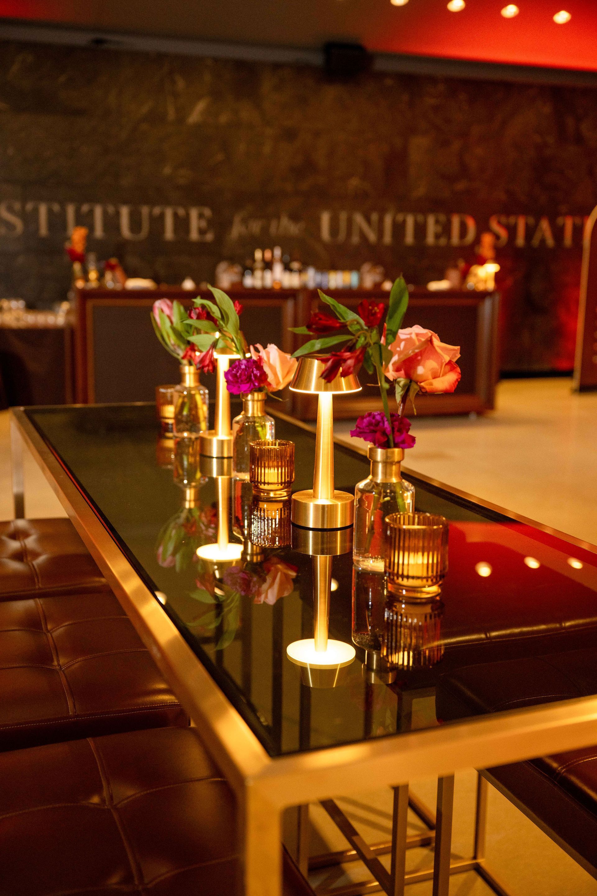 Long table with flowers in vases, gold accents, and a bar in the background.