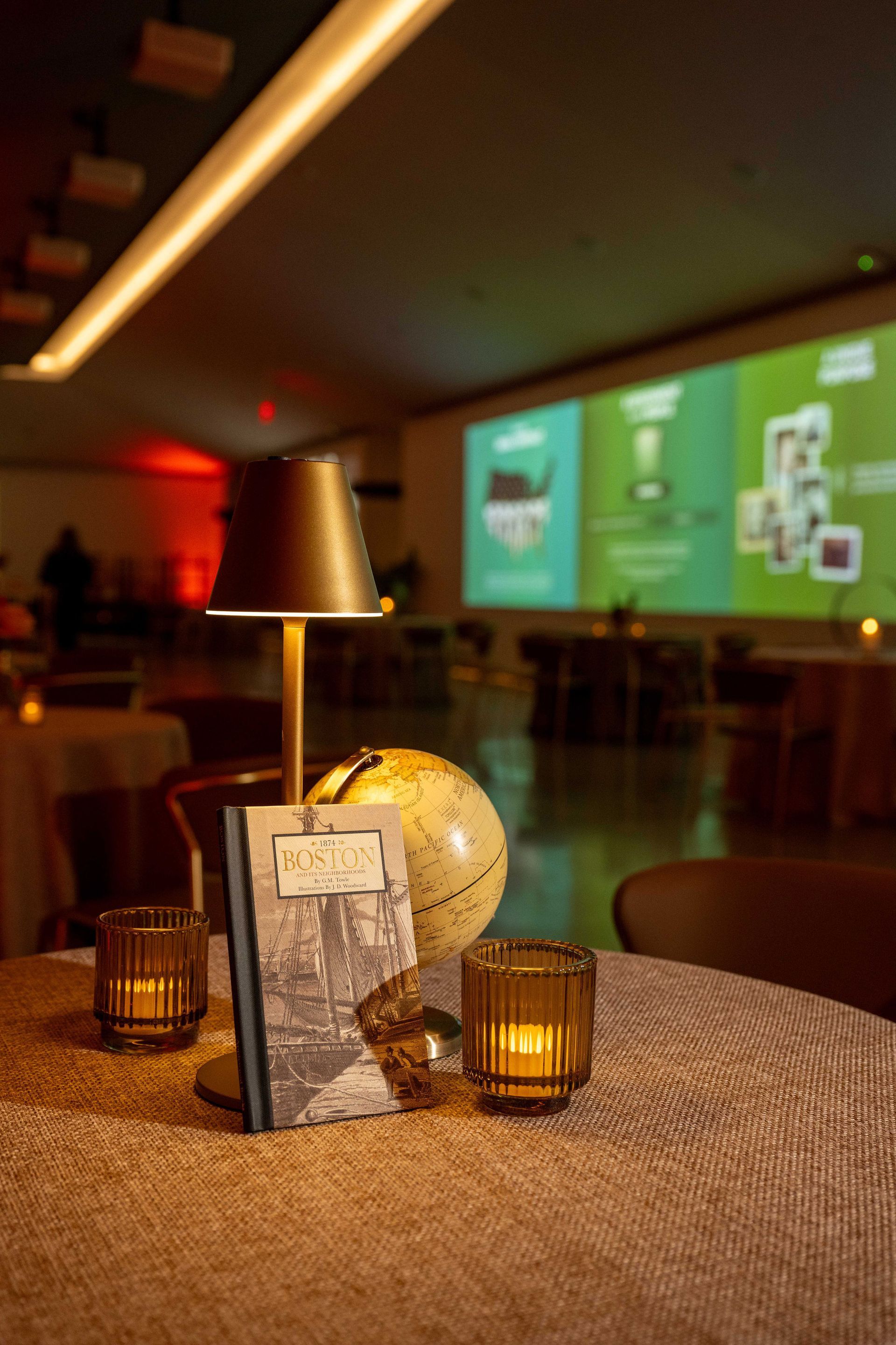 A table centerpiece with a book, globe, lamp, and candles. Blurred background with a screen and tables.