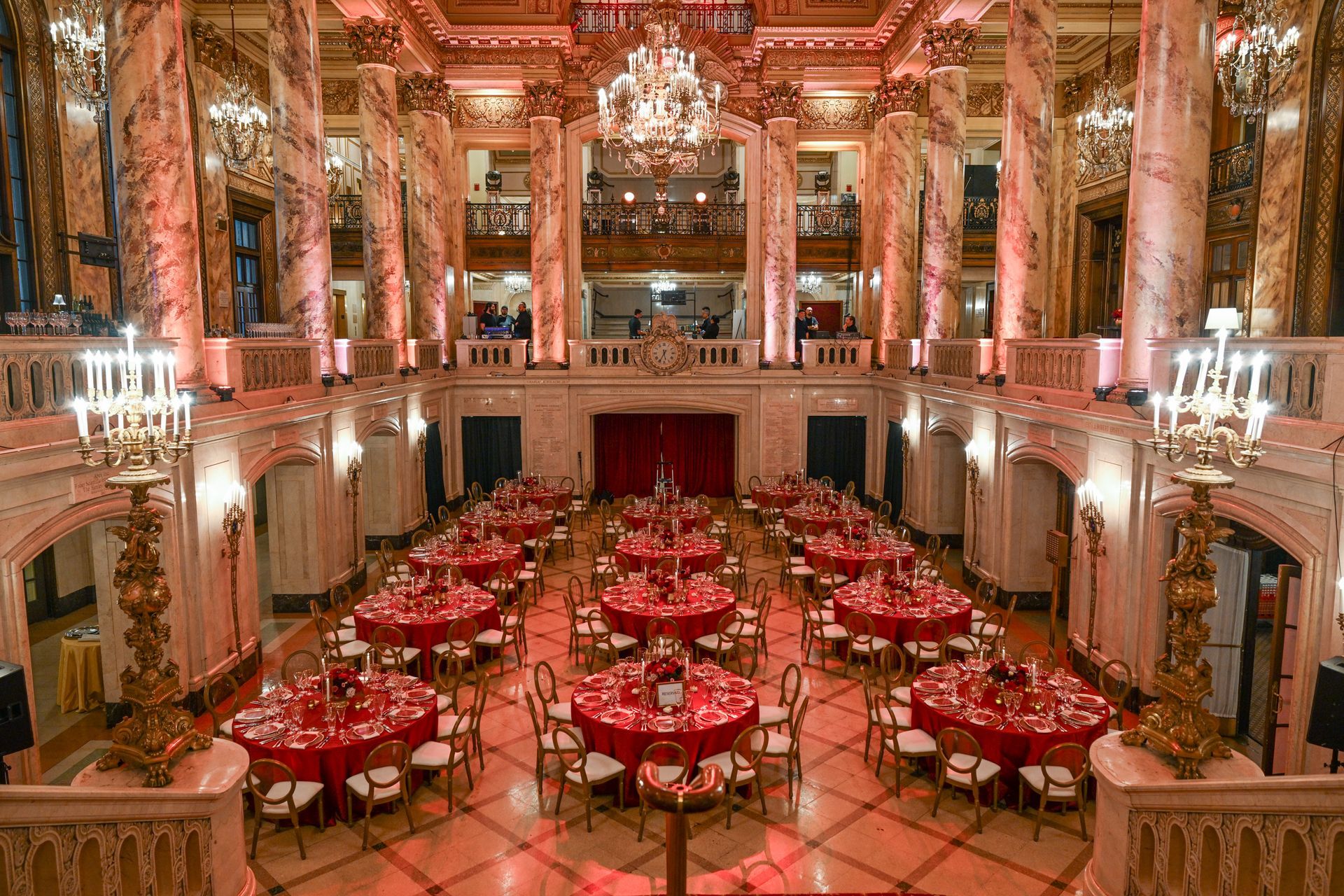 Elegant ballroom set for a formal event; red tables and decor, marble columns, grand chandeliers.