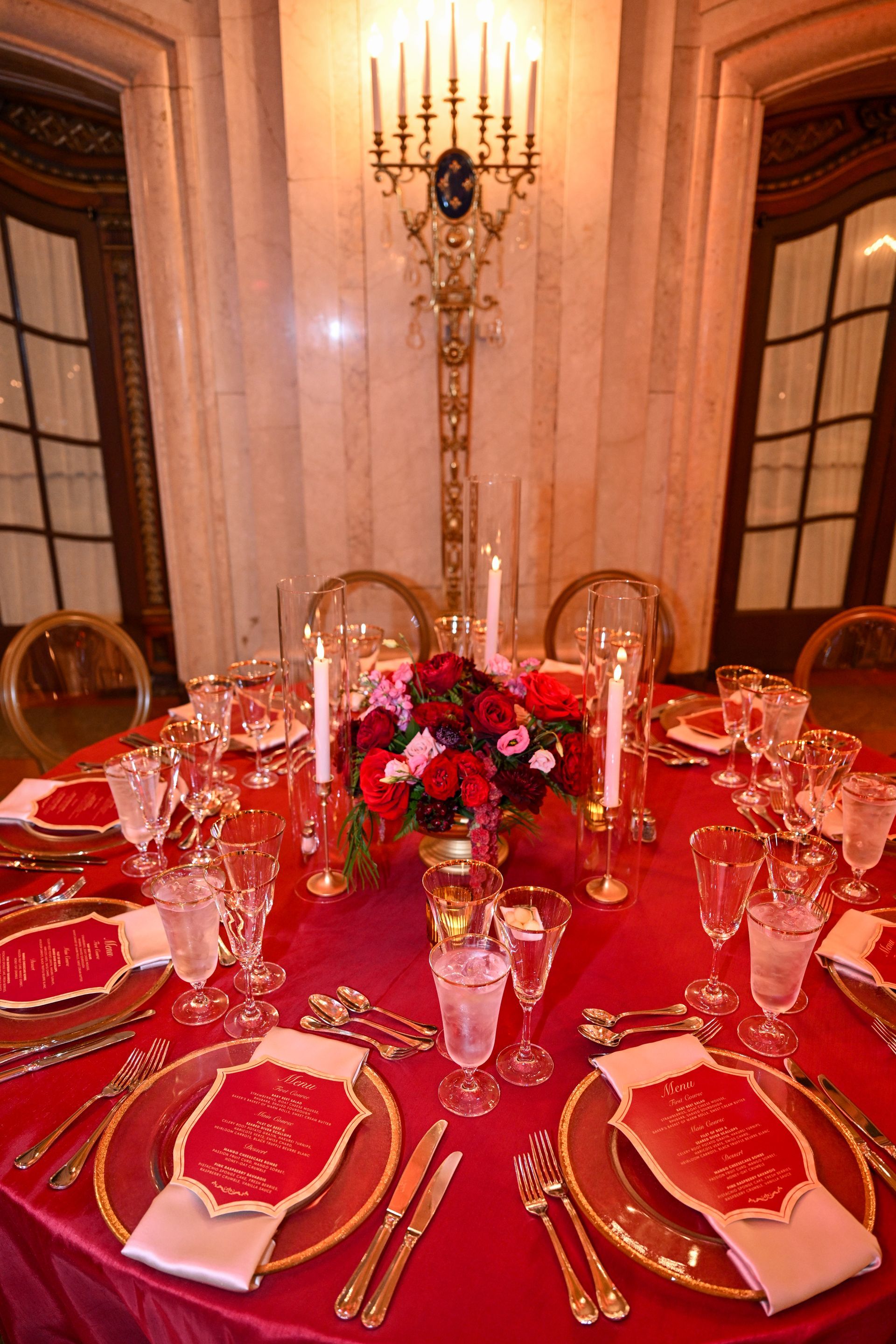 Formal dining table set with red tablecloth, gold-rimmed plates, silverware, and floral centerpiece, lit by candles.
