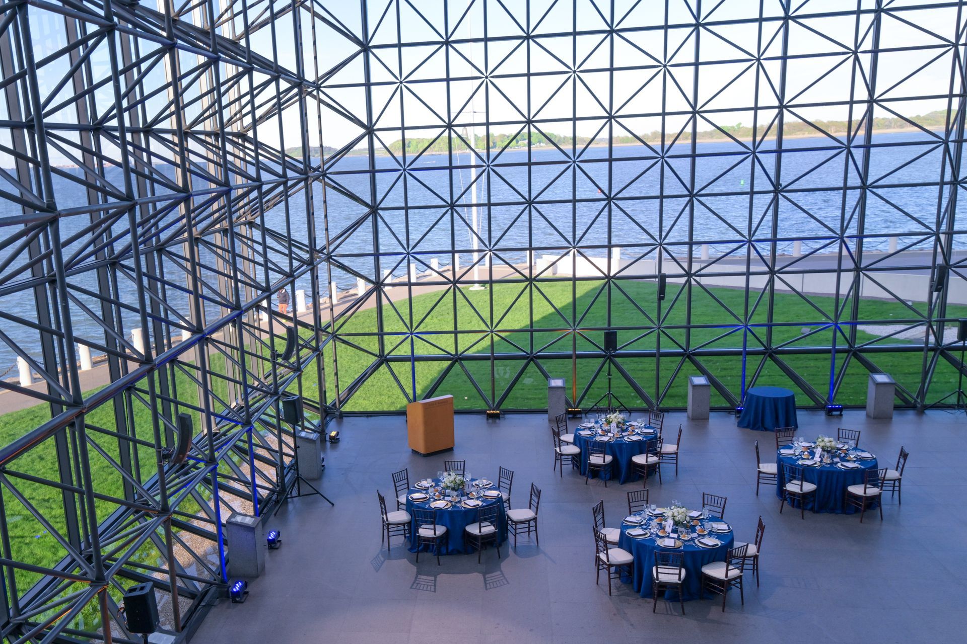 Large event space with tables, overlooking water. Blue linens, geometric glass facade.