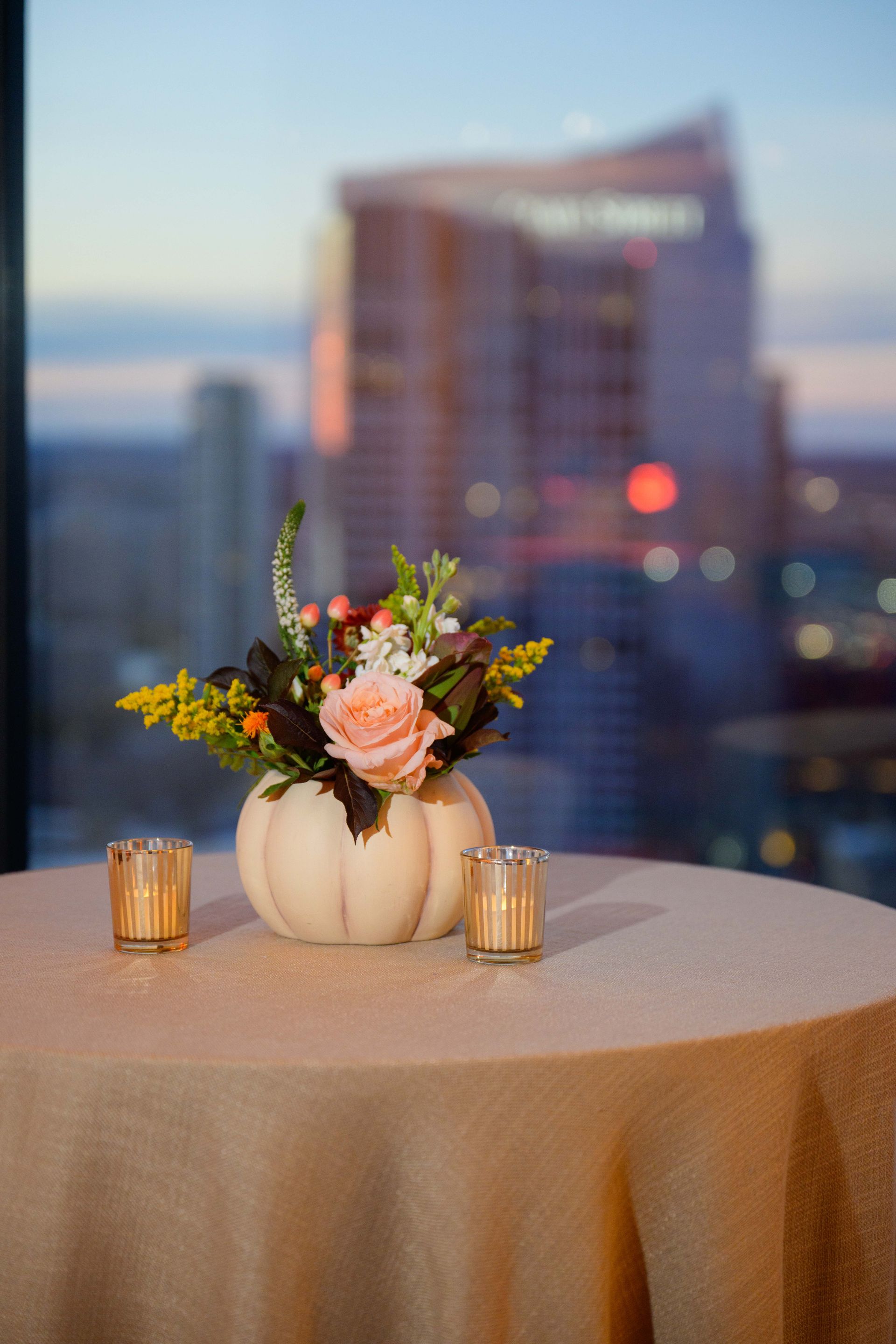 Table set for a party with pumpkin flower arrangement and candle, cityscape background at sunset.