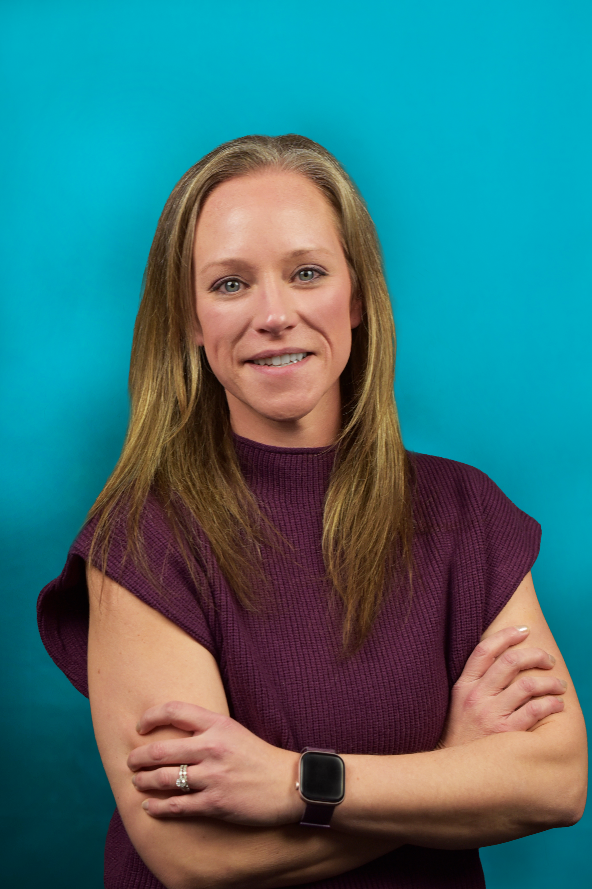 Woman with crossed arms smiles; wears a purple sweater, smart watch, and stands against a blue backdrop.