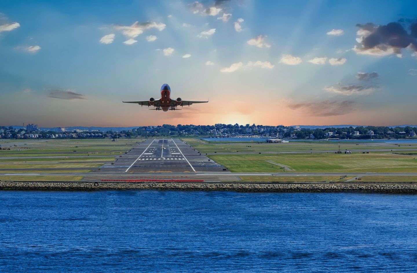 Airplane taking off over water with the runway in view, city skyline in the distance, sunset sky.