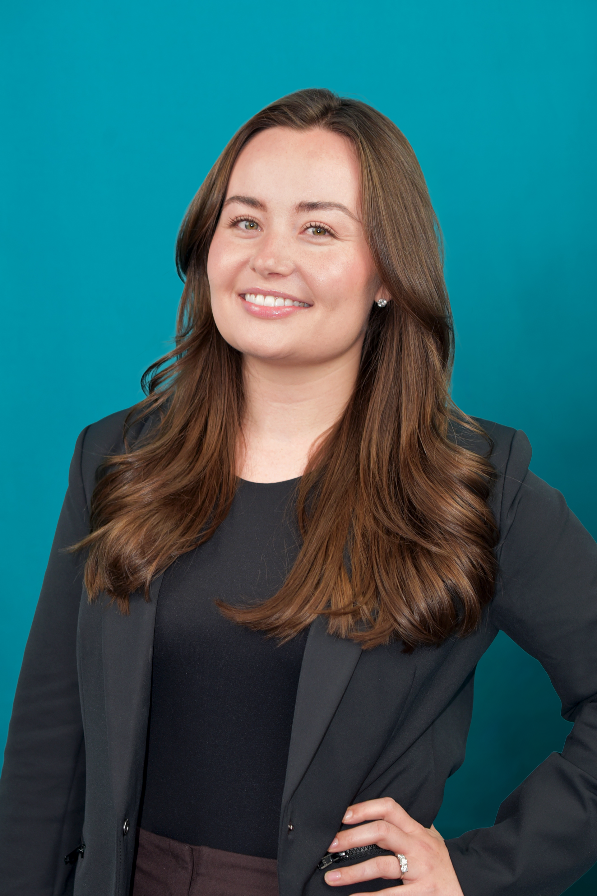 Woman in a black blazer and top smiling with her hand on her hip, turquoise background.