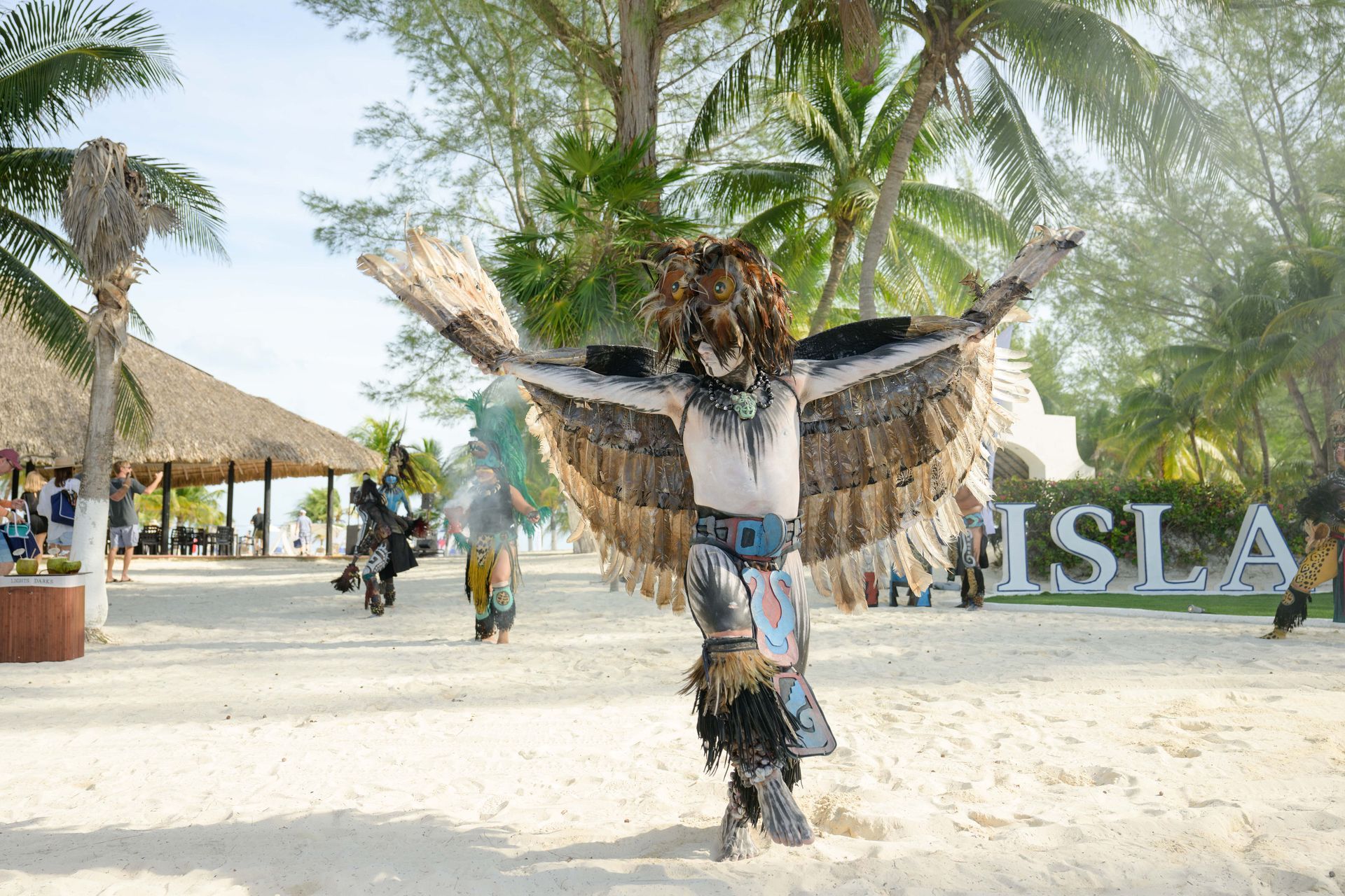 Dancers in feathered costumes perform on a sandy beach near 