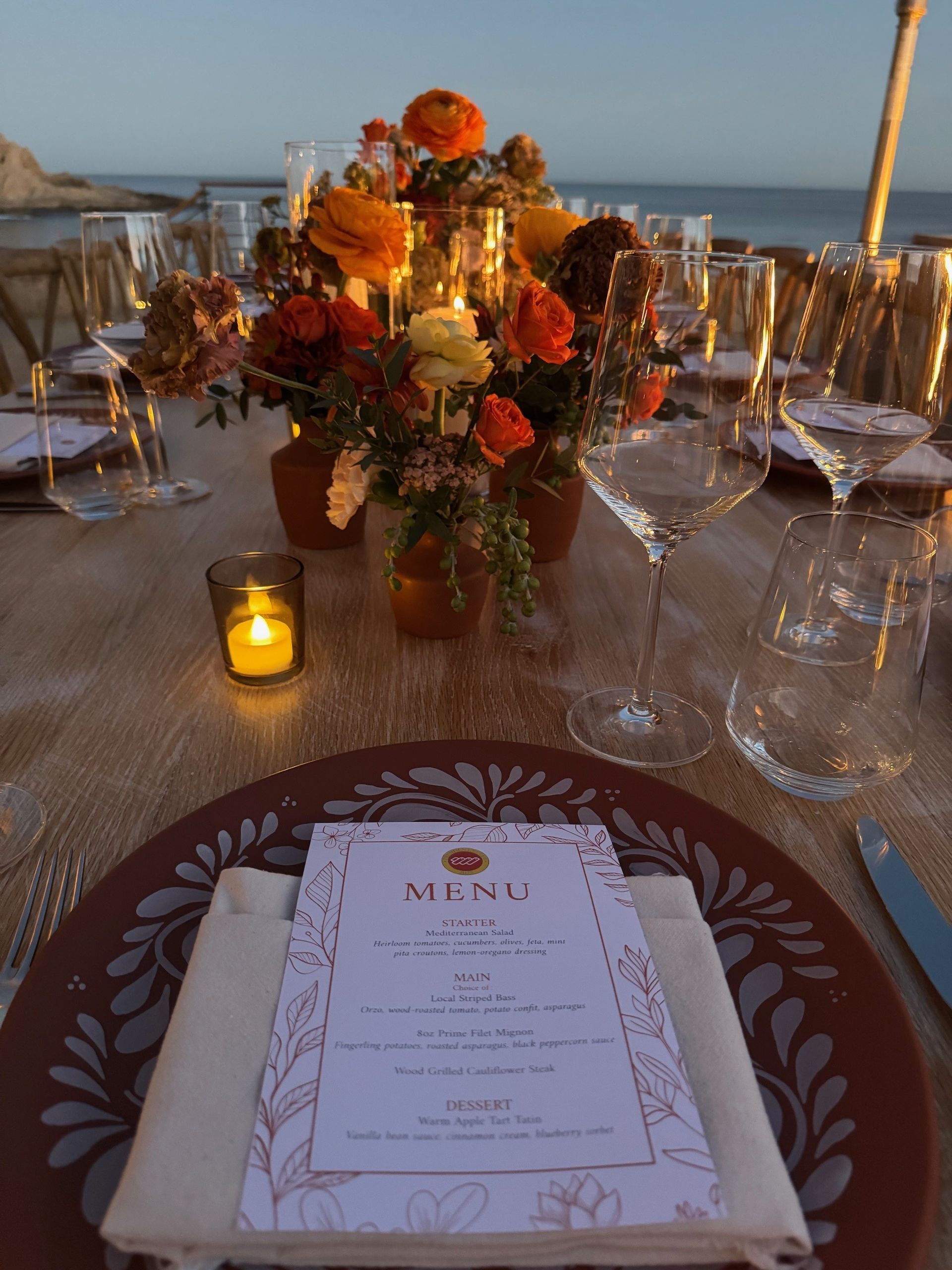 Elegant table setting with floral centerpiece, lit candles, and menu card, overlooking the ocean at sunset.