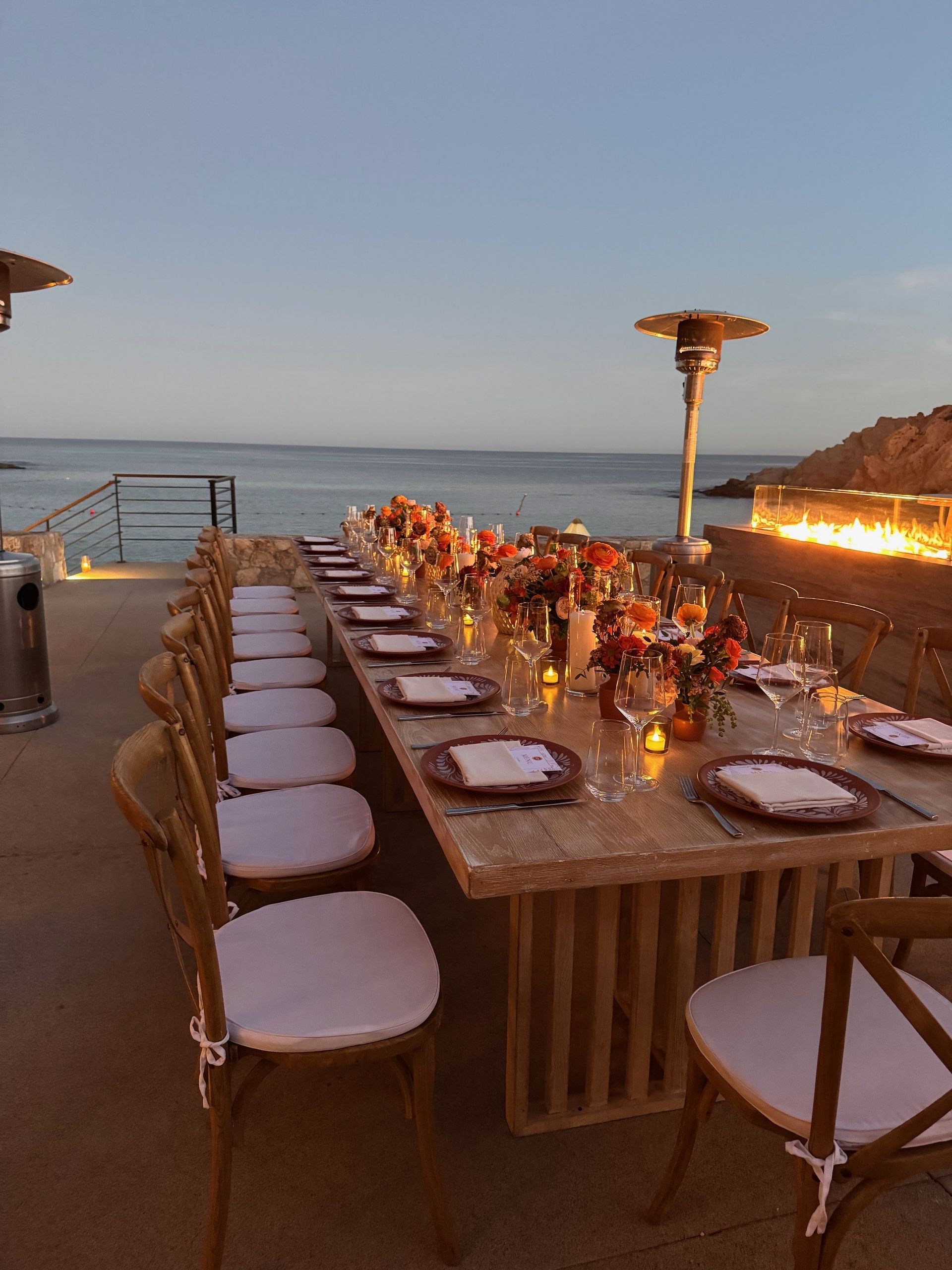 Long outdoor table set for a dinner party near the ocean at dusk.